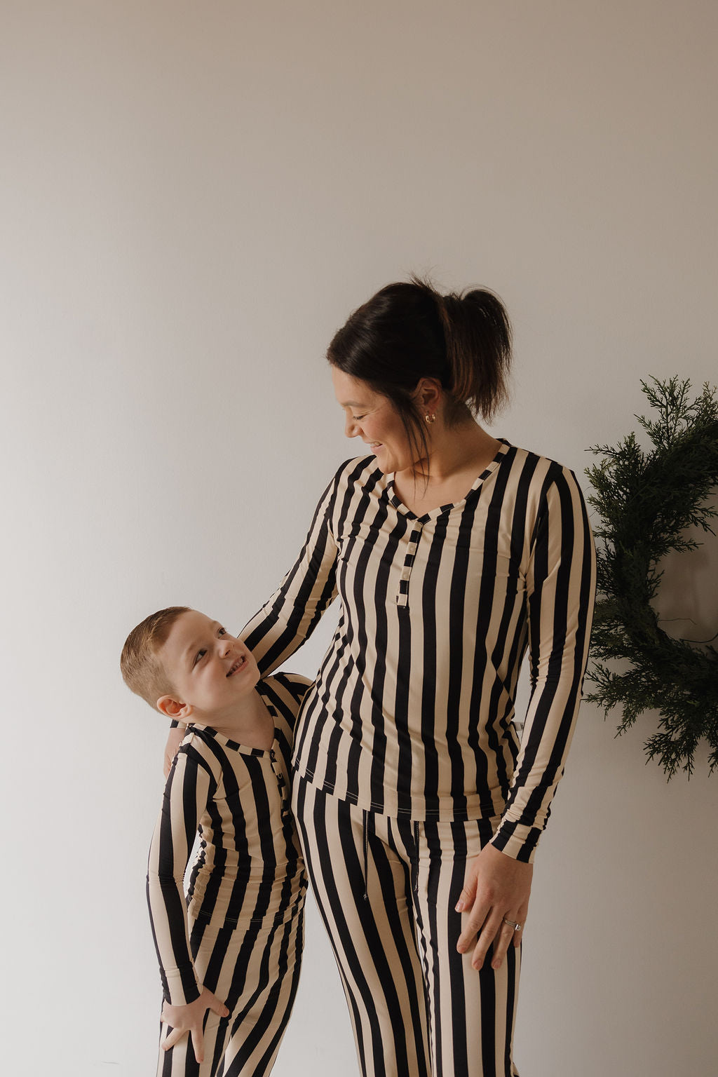 A woman in the forever french baby Women's Long Sleeve Bamboo Pajama in Midnight Stripe stands closely with a smiling young boy; both wear matching black and beige striped outfits against a plain background with a green wreath.