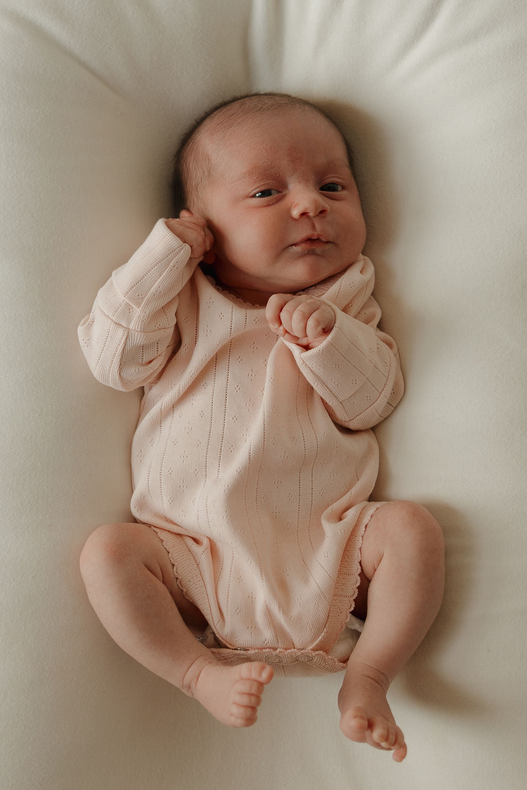 A newborn baby lies on a soft white blanket, dressed in the forever french baby Long Sleeve Snapsuit | Ballerina. The light pink pointelle cotton snapsuit features long sleeves, as the baby gazes calmly at the camera with one hand near their face and their legs gently bent.