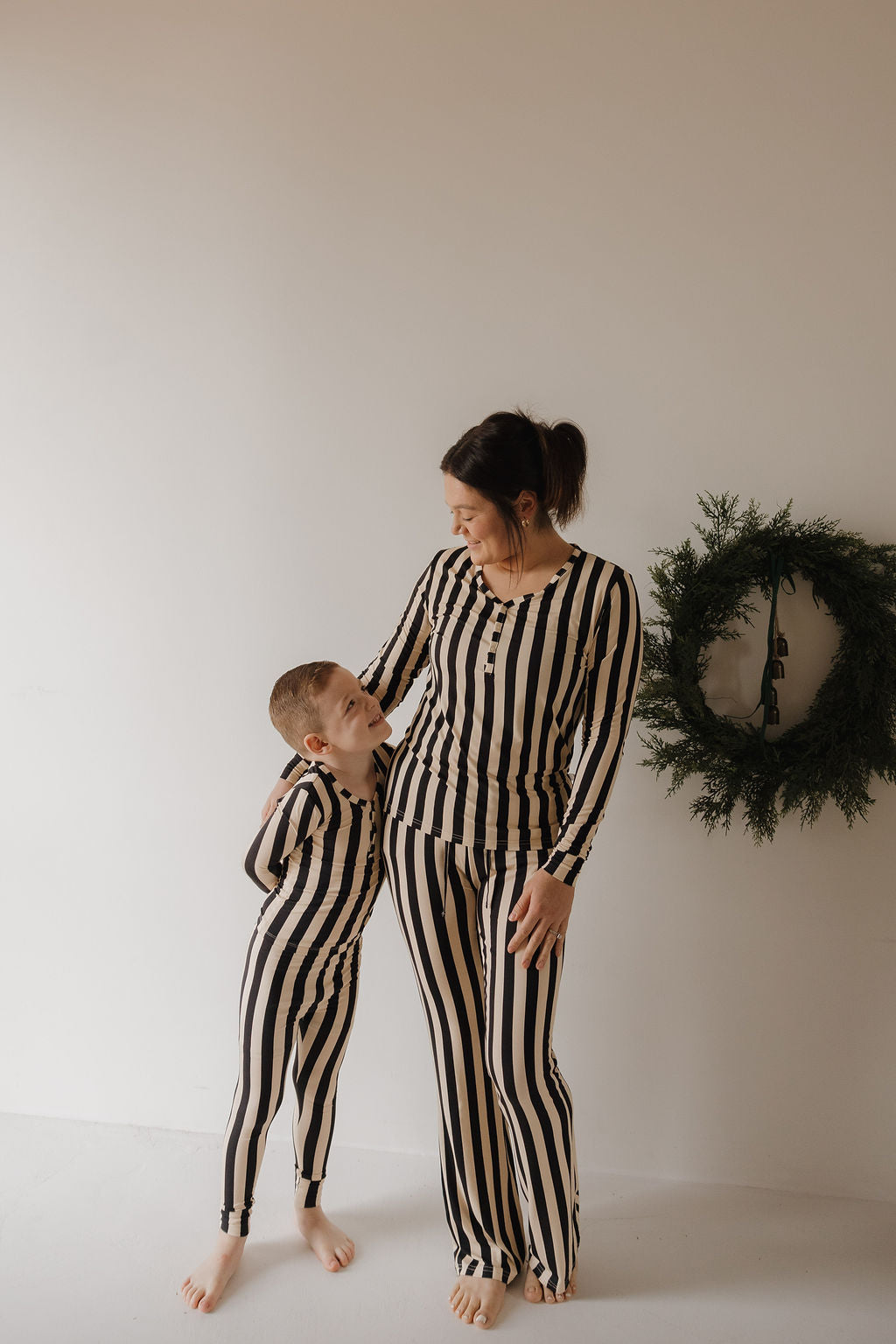 A woman and child smile at each other in a minimalist room, wearing Minty Cherries Bamboo Two Piece Pajamas in Midnight Stripe—breathable black and beige sleepwear perfect for matching moments.