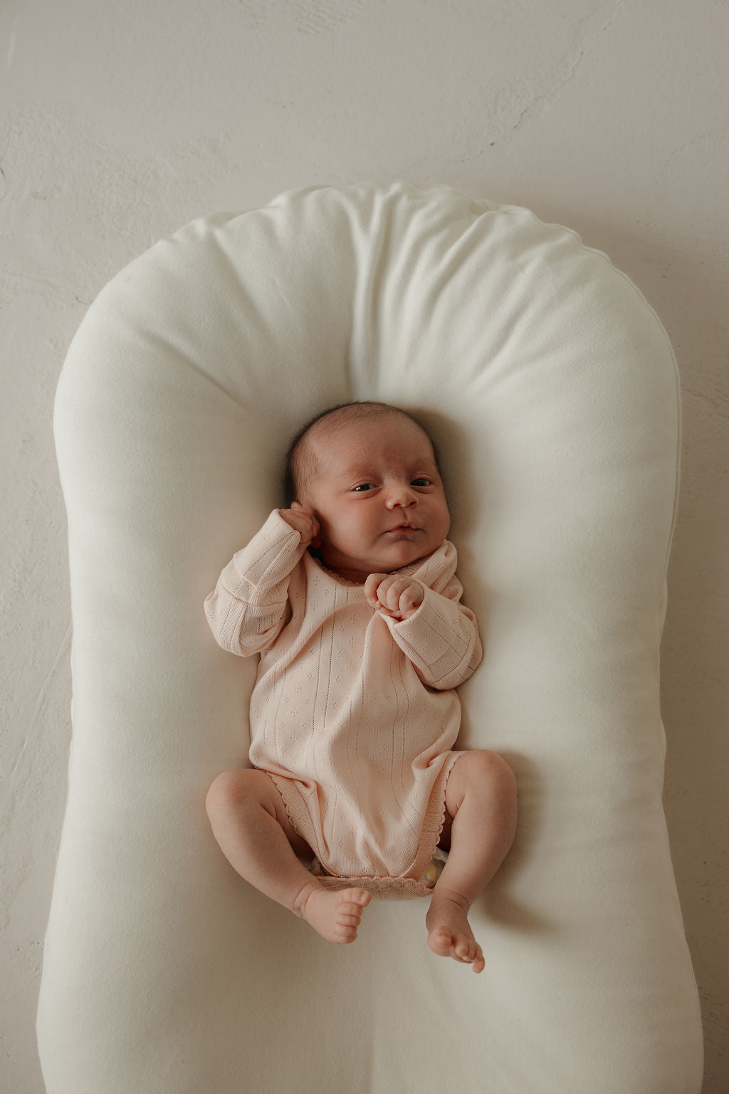 A newborn baby wearing the forever french baby Long Sleeve Snapsuit in Ballerina rests on a soft, white, oval-shaped cushion, gazing up with a relaxed expression.