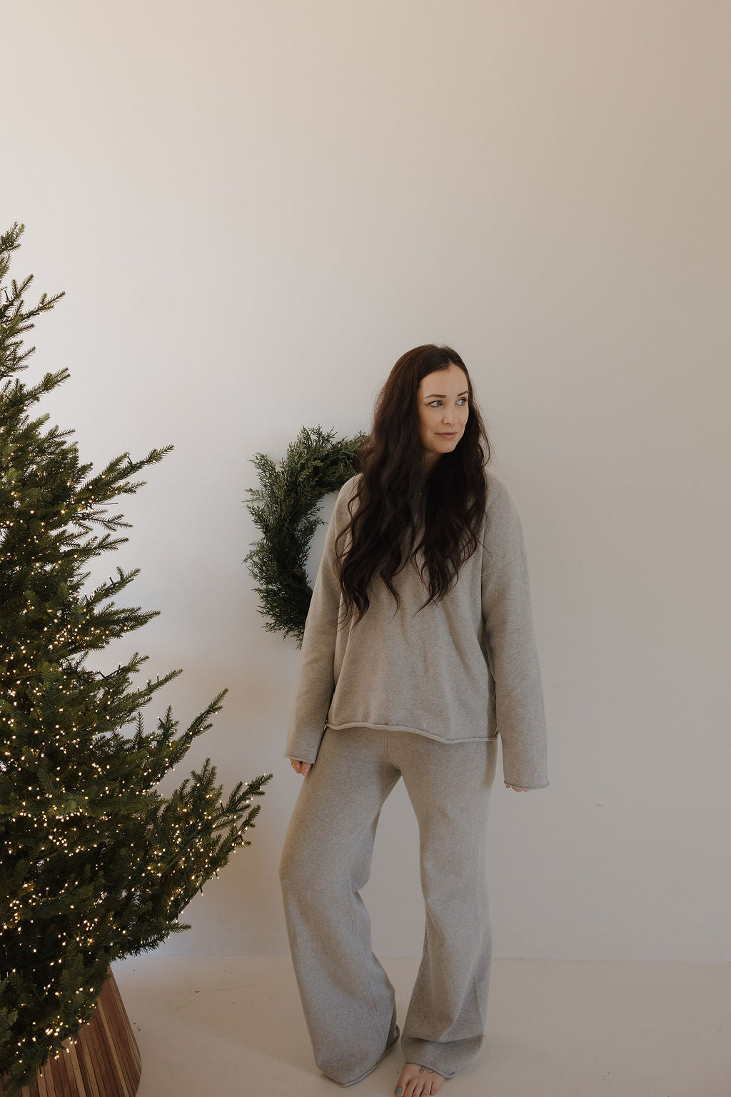 A woman with long dark hair stands barefoot by a Christmas tree and green wreath, wearing the forever french baby Women's Knit Pant Set in grey.