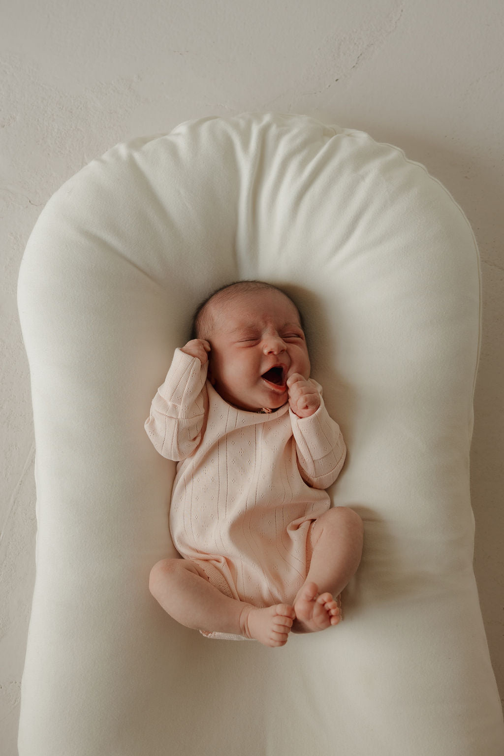 A newborn baby wearing the forever french baby Long Sleeve Snapsuit | Ballerina in light pink pointelle cotton lies on a soft, white cushion, yawning with eyes closed and one fist near their mouth.
