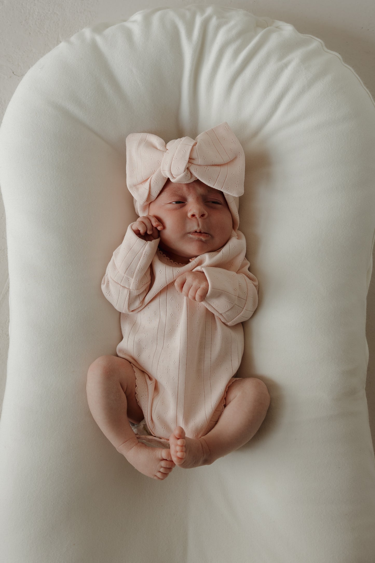 A baby wearing the forever french baby Pointelle Head Wrap in Ballerina and a light pink pointelle cotton outfit lies on a soft, white cushion, gazing upward with a relaxed expression.