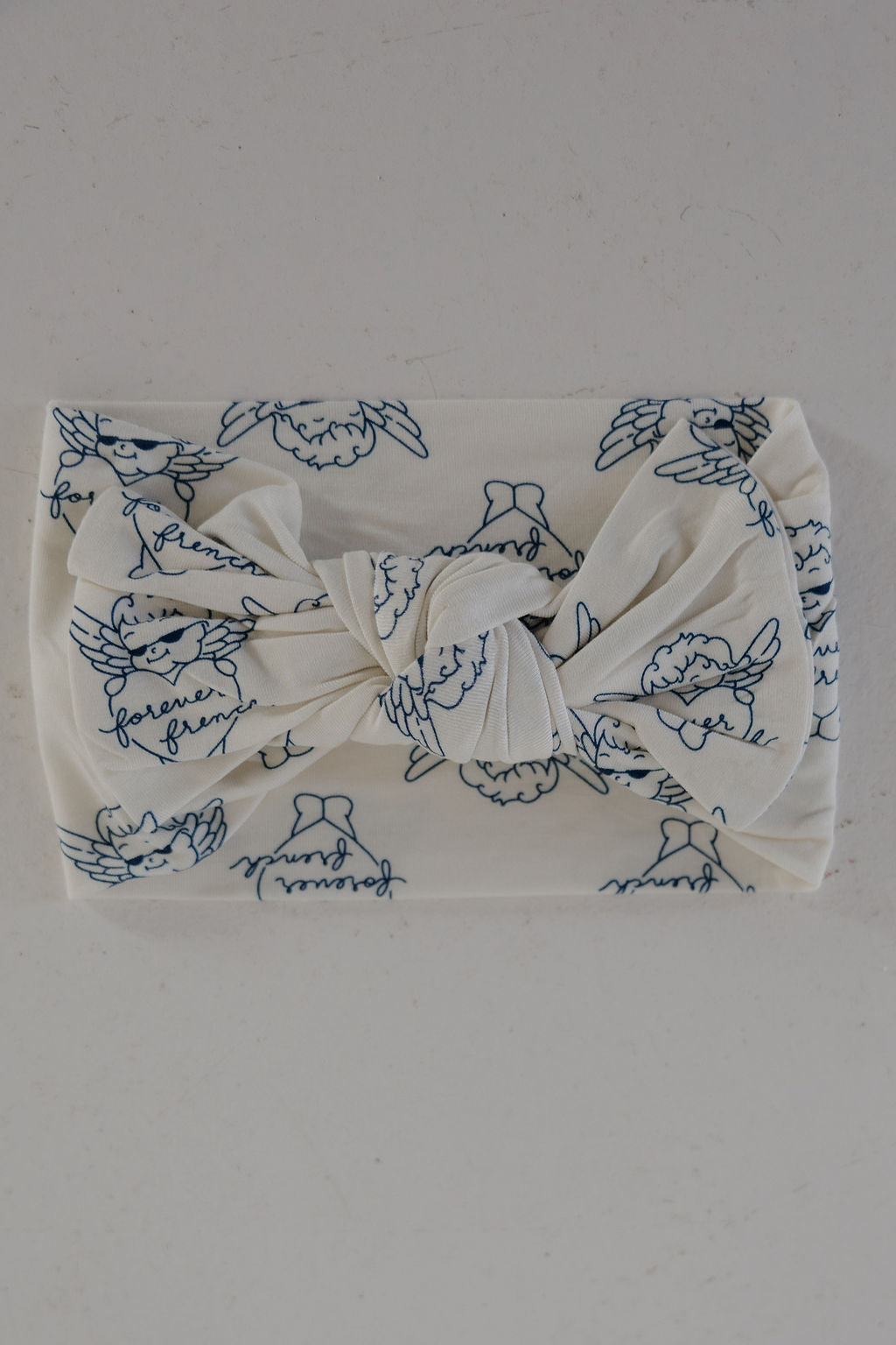The Bamboo Head Wrap | ff Los Angeles by forever french baby is a white baby bow headband with a front knot and blue cherub, bell, and cursive pattern, displayed on a light gray surface.