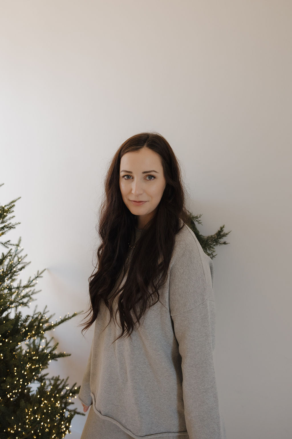 A woman with long dark hair wears the forever french baby Women's Knit Pant Set in grey, standing indoors near a decorated Christmas tree with white lights against a plain light-colored wall.