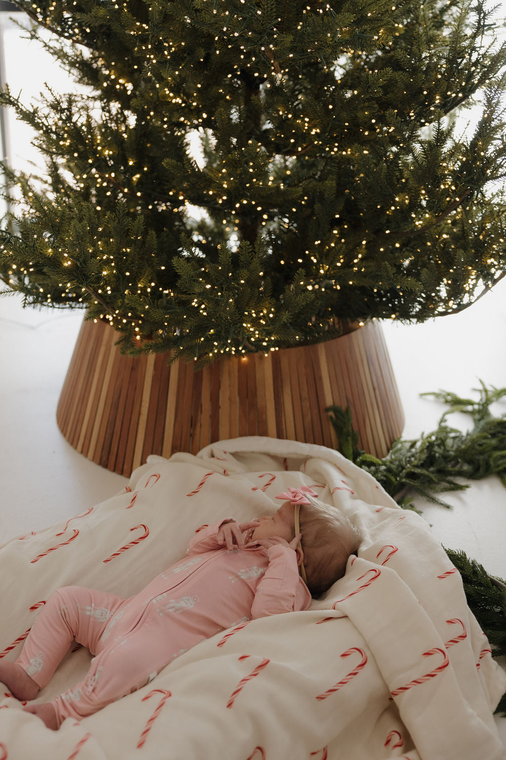 A baby in forever french baby's Bamboo Zip Pajamas | Nutcracker lies on a white blanket with candy canes, gazing up at a Christmas tree lit with warm white lights.
