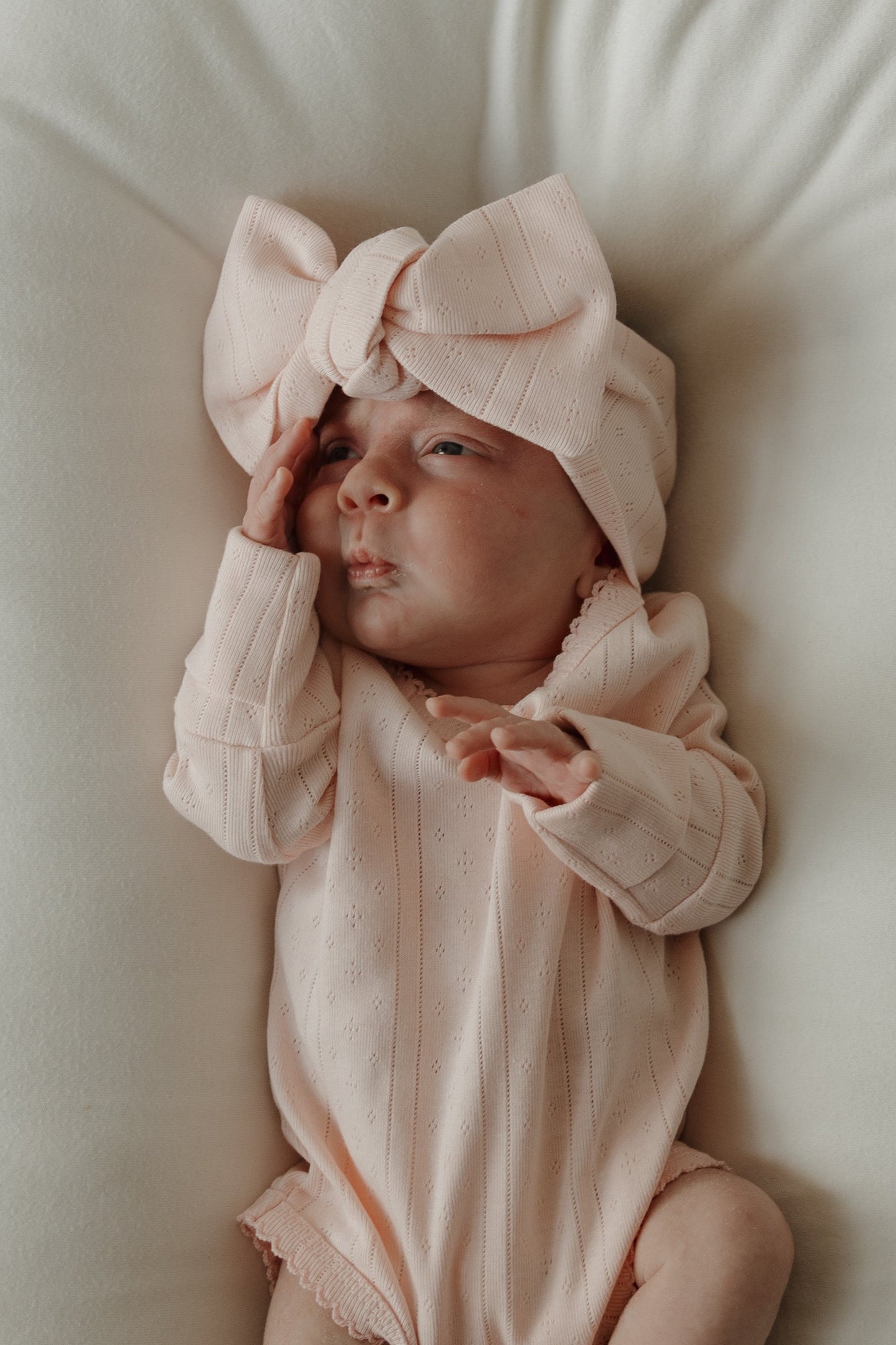 A baby wearing a light pink pointelle cotton onesie and the forever french baby Pointelle Head Wrap in Ballerina lies on a white blanket, with one hand touching their face and the other resting on their chest.
