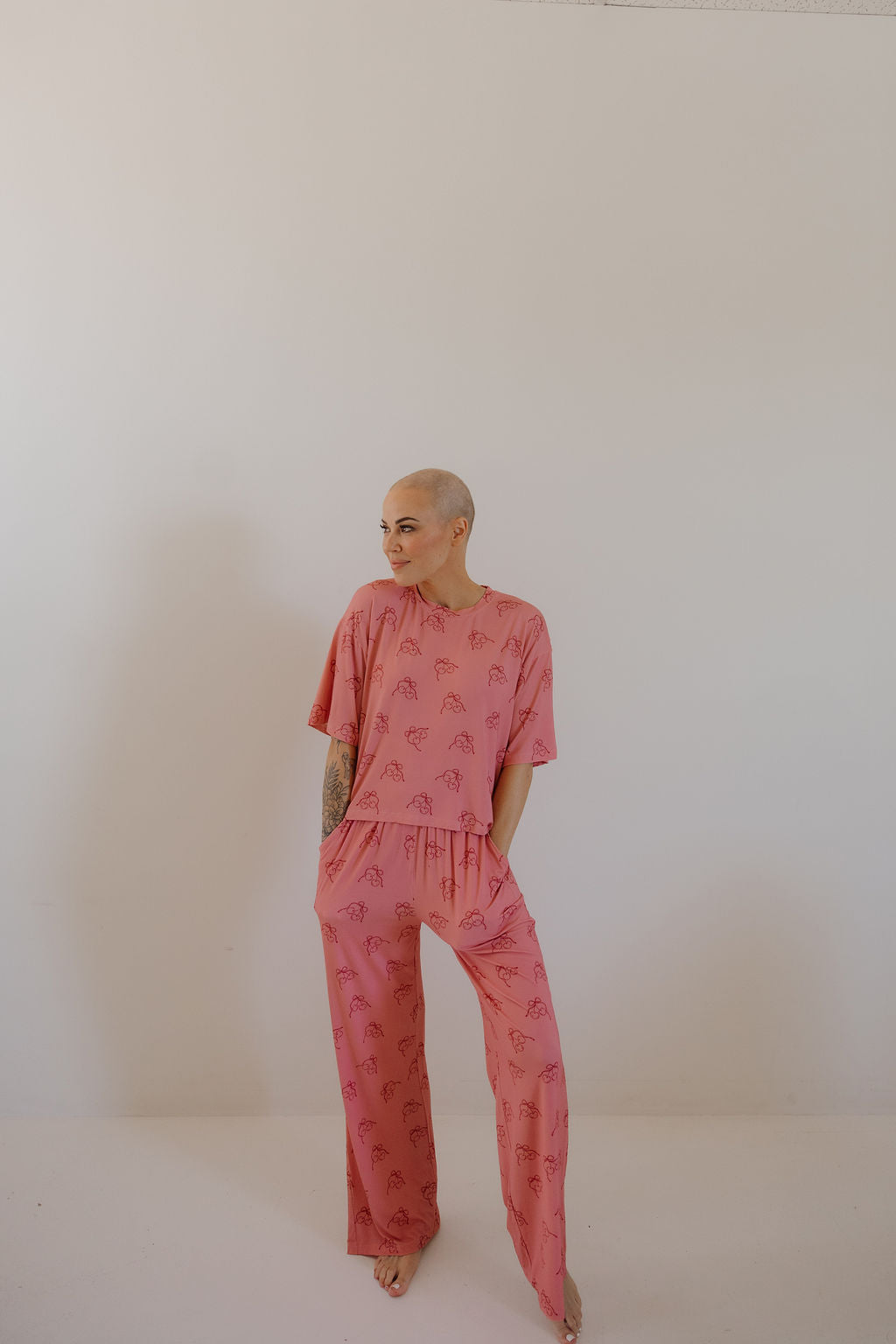 A person with a shaved head stands barefoot against a plain white background, wearing the Minty Cherries Women's Boxy Tee Lounge Set in Iconic Cherry, hands in pockets and looking to the side.