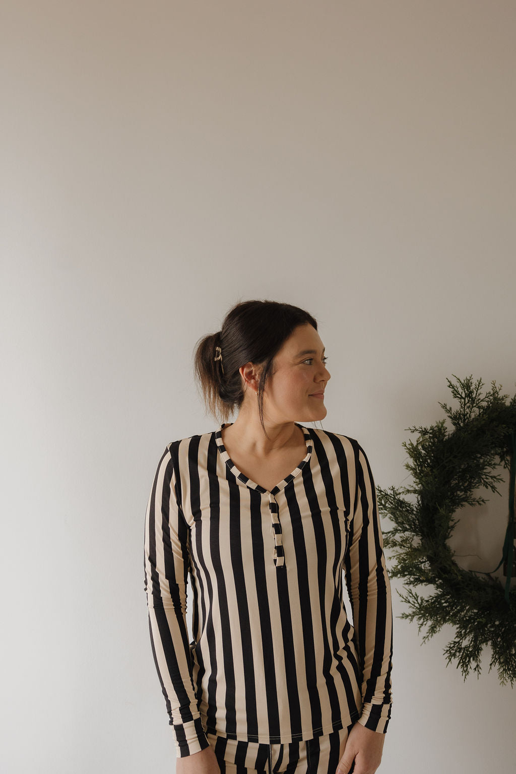 A woman wearing the forever french baby Women's Long Sleeve Bamboo Pajama in Midnight Stripe stands by a plain wall, looking left. A green wreath hangs on the right side of the image.