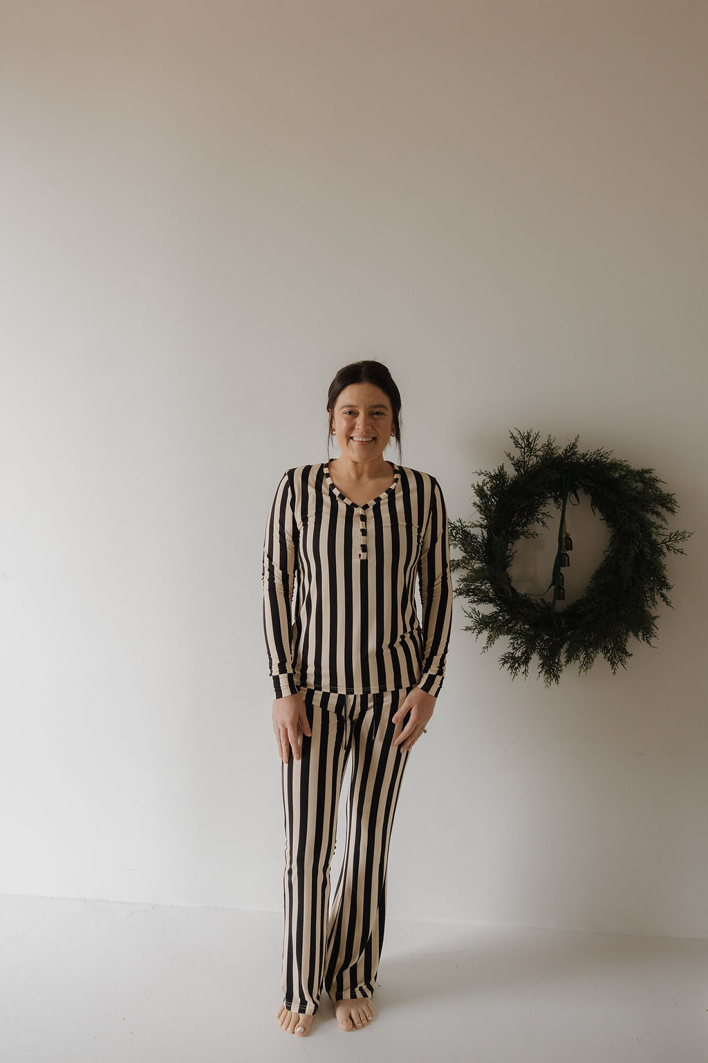 A woman smiles at the camera barefoot indoors, wearing the forever french baby Women's Long Sleeve Bamboo Pajama in Midnight Stripe. A green wreath on a white wall highlights her stylish and breathable bamboo sleepwear.