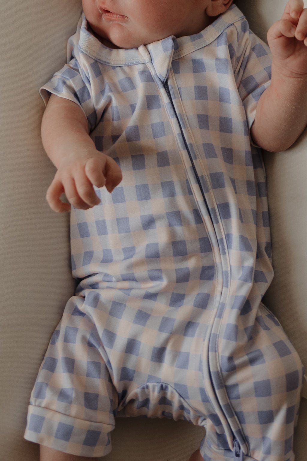 A baby wearing the forever french baby Shortie Bamboo Zip | Marilyn—featuring a classic blue and white checkered design—lies on a light-colored surface, one hand slightly raised and only part of their face visible. This hypoallergenic romper offers both comfort and style for your little one.