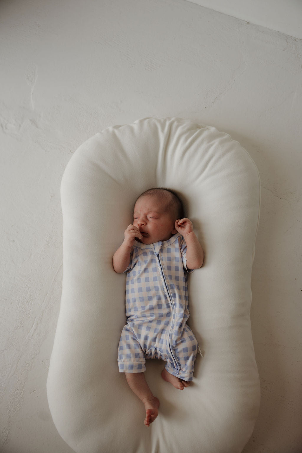 A newborn baby in forever french baby’s Shortie Bamboo Zip | Marilyn—blue and white checkered, with eyes closed and hand near mouth—rests on a white cushioned lounger against a light background, perfectly cozy in hypoallergenic clothing designed for delicate skin.