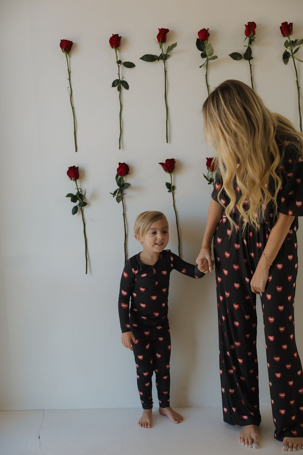 A woman and a child, barefoot and holding hands, wear matching forever french baby Bamboo Two Piece Pajamas | Love Notes – Charcoal, featuring red lips, in front of a white wall decorated with vertical red roses.