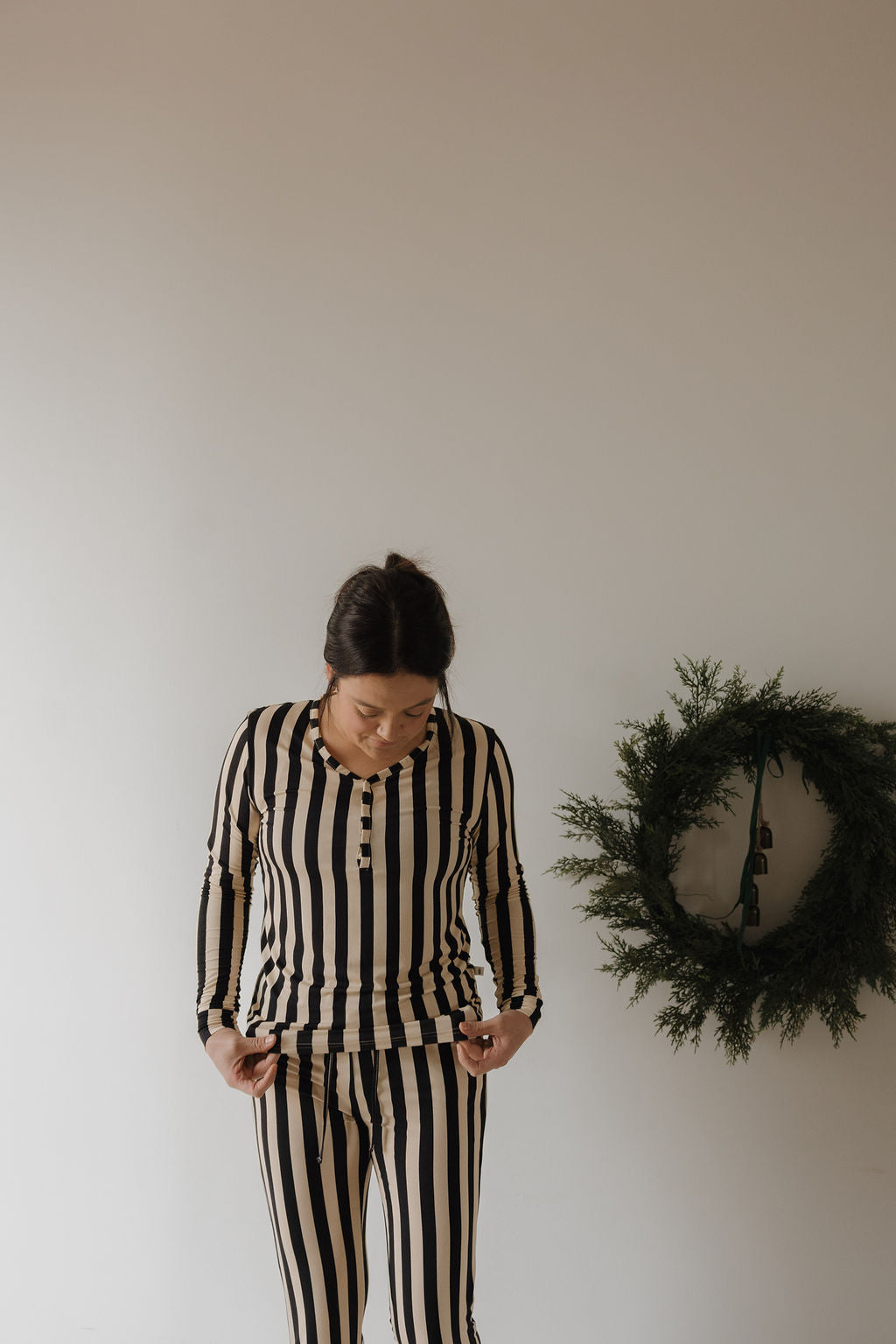 A woman in the forever french baby Women's Long Sleeve Bamboo Pajama in Midnight Stripe stands by a plain wall, looking down. A green wreath hangs on the wall to her right.