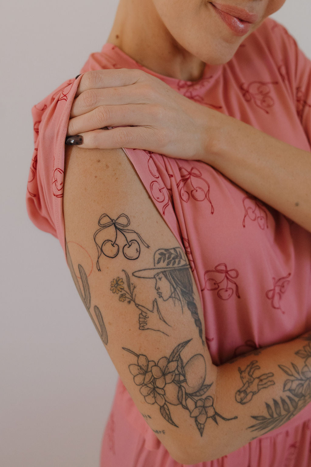 A person in the Minty Cherries Women's Boxy Tee Lounge Set | Iconic Cherry holds their shoulder, showing cherry and botanical tattoos on their arm. Their face is partly visible with a slight smile.