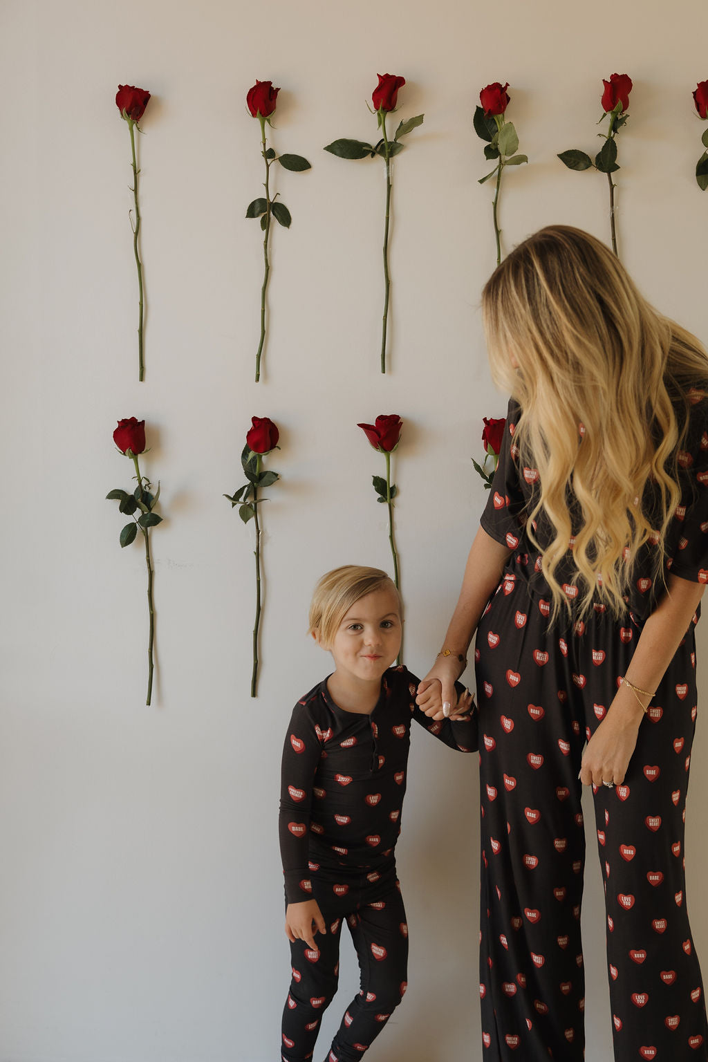 A woman and child wear matching forever french baby Bamboo Two Piece Pajamas | Love Notes – Charcoal, both featuring a red lips print, as they stand before a white wall decorated with rows of red roses.