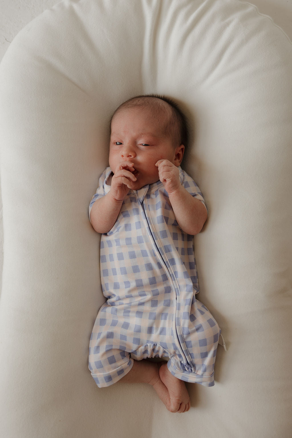A newborn baby wearing the forever french baby Shortie Bamboo Zip | Marilyn—a blue and white checkered romper—lies on a soft, white cushion, gazing upward with hands near the mouth and legs slightly bent. The hypoallergenic fabric of this romper provides ultimate comfort for delicate skin.