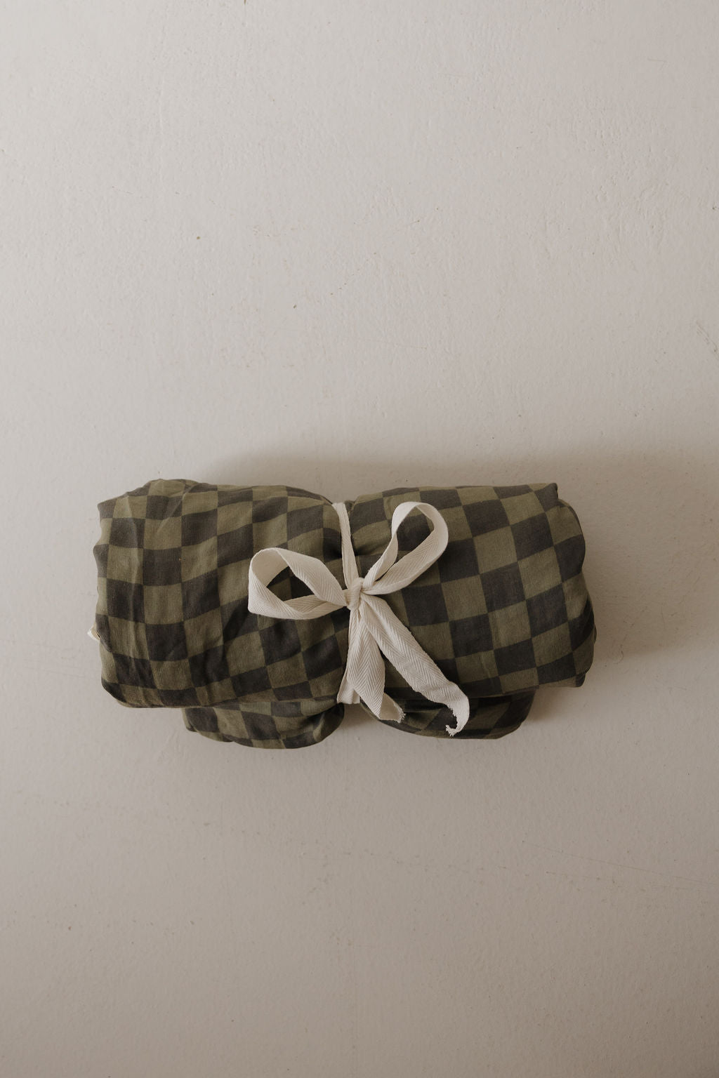 The Muslin Quilt | Olive Checkerboard by forever french baby, in green and black, is neatly rolled with a white ribbon against an off-white background—an ideal choice for stylish nursery bedding.