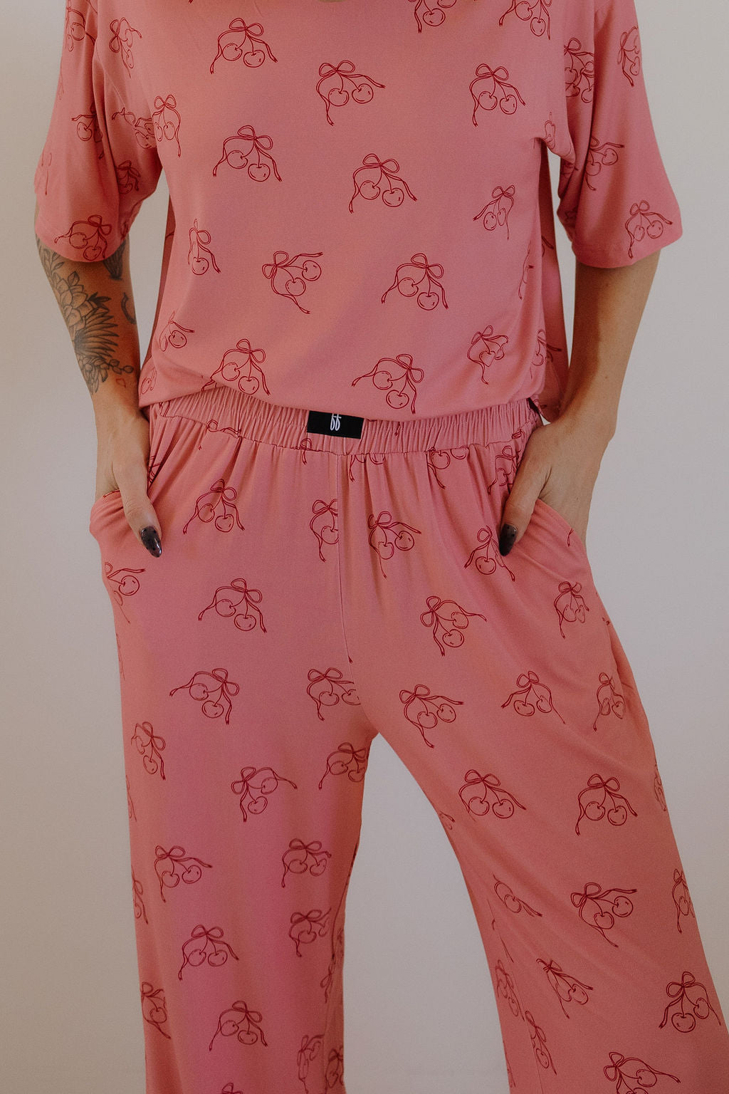 A person wears Minty Cherries' Women's Boxy Tee Lounge Set in Iconic Cherry, featuring a pink set with a red abstract face print. The short-sleeve top and elastic-waist pants are shown from the neck down, hands in pockets, against a light background.