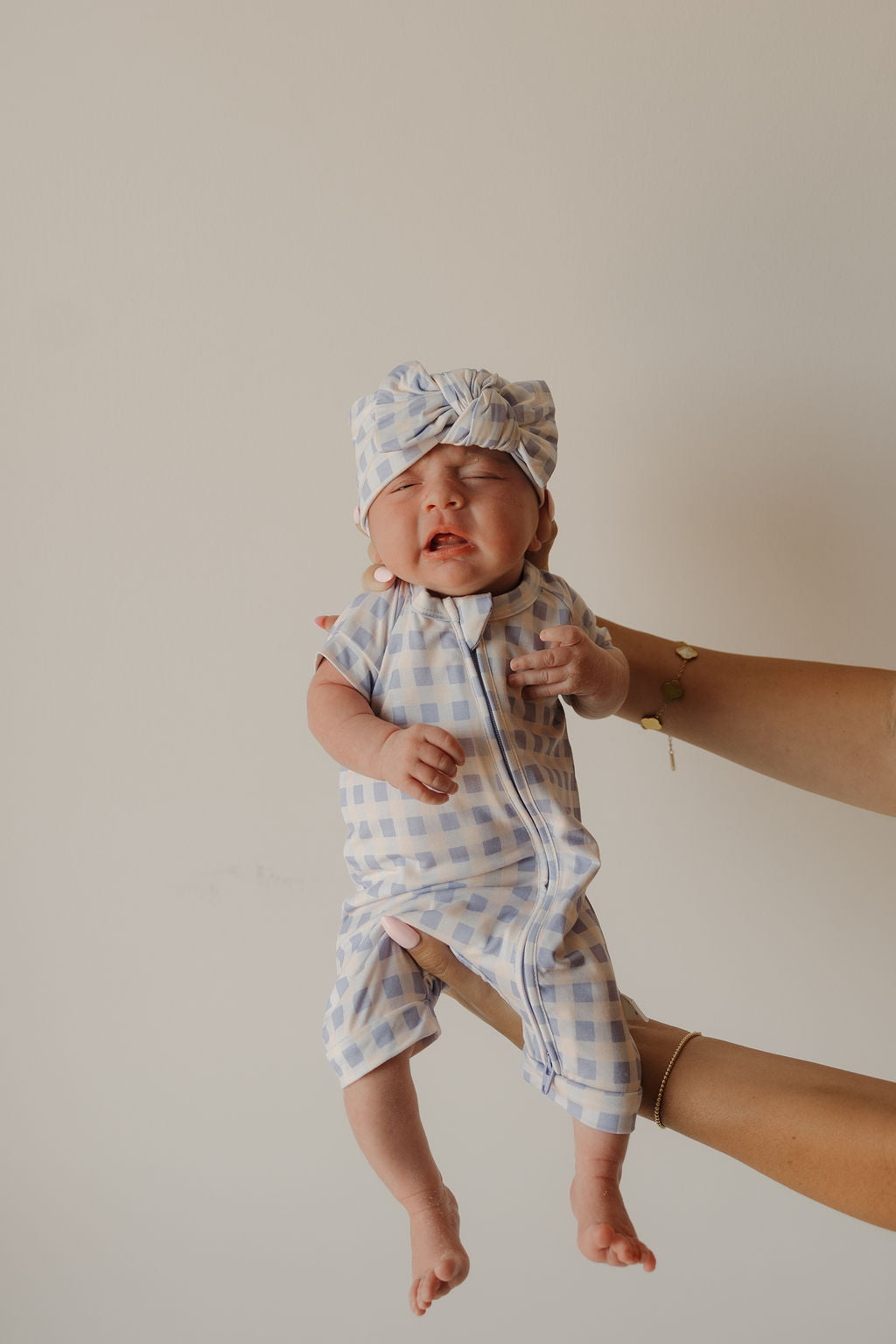 An adult hand holds up a crying baby dressed in the forever french baby Shortie Bamboo Zip | Marilyn—a hypoallergenic blue and white checkered outfit with a matching hat—against a plain light background.