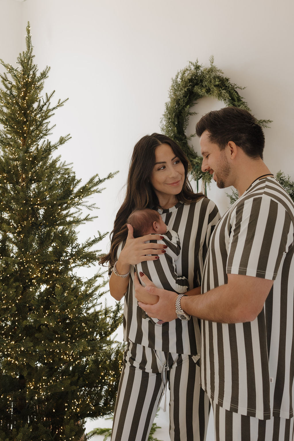 A woman in forever french baby Short Sleeve Women's Bamboo Pajamas in Fireside Stripe stands with her matching family by a decorated Christmas tree, sharing warm smiles.