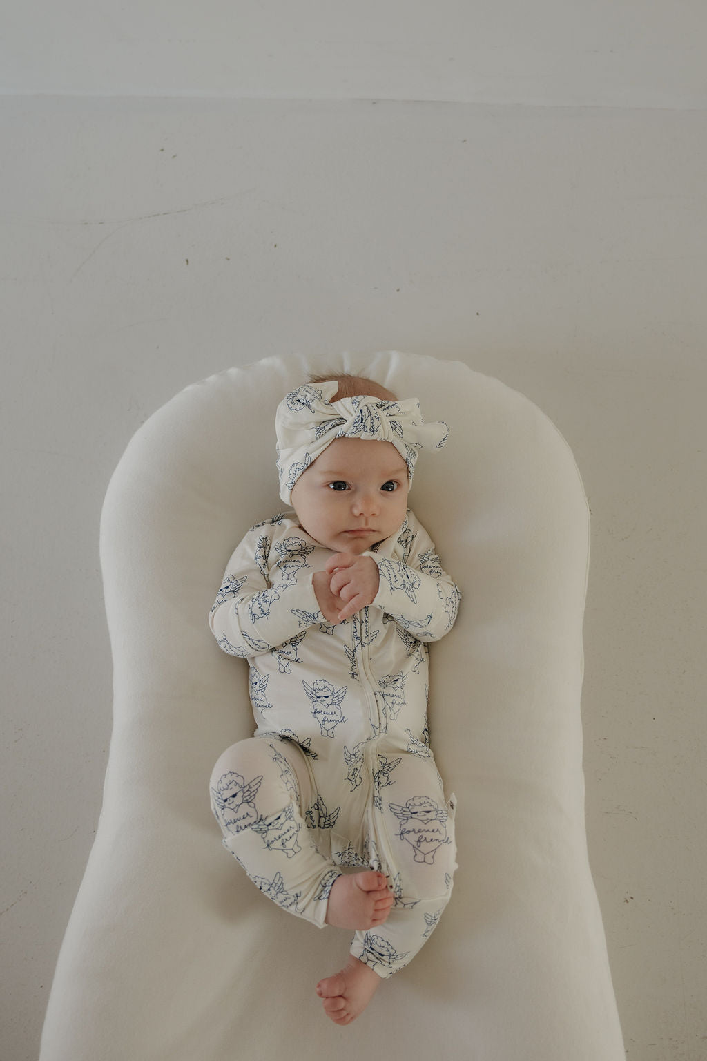 A baby in a forever french baby onesie and a Bamboo Head Wrap | ff Los Angeles from forever french baby lies on a white cushioned surface, gazing upward with hands clasped together.