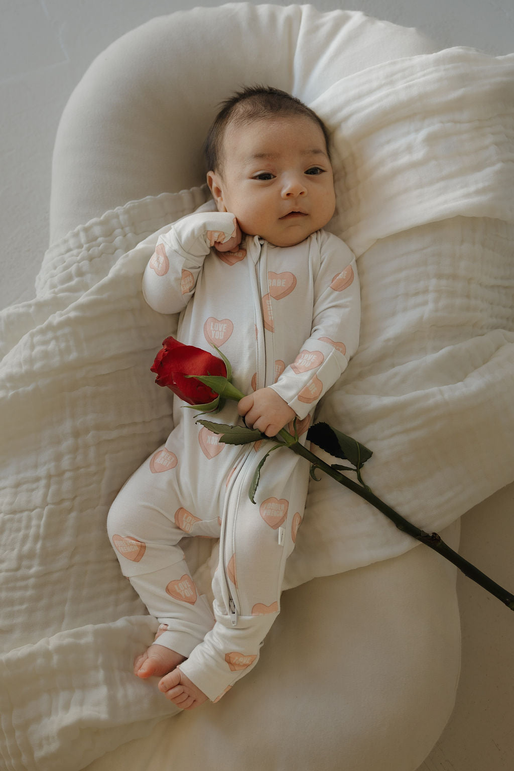 A baby in forever french baby's Bamboo Zip Pajamas | Love Notes - Grey rests calmly on a soft cushion, holding a red rose, with one arm bent by their head.