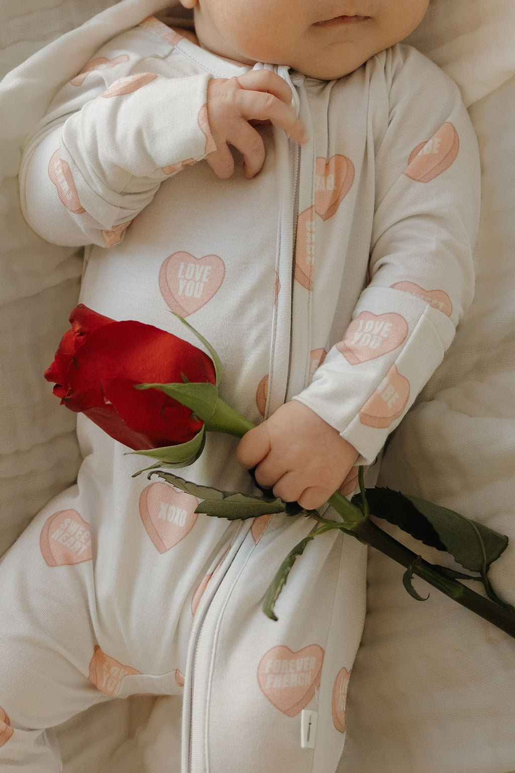 A baby in forever french baby's Bamboo Zip Pajamas | Love Notes - Grey holds a red rose while lying on a soft, cream-colored blanket.