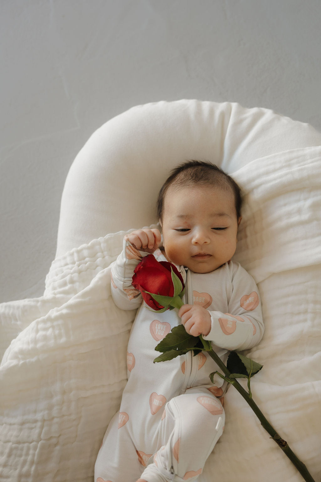A baby in forever french baby's Bamboo Zip Pajamas | Love Notes - Grey lies peacefully on a soft white cushion, holding a red rose and partially covered by a blanket.