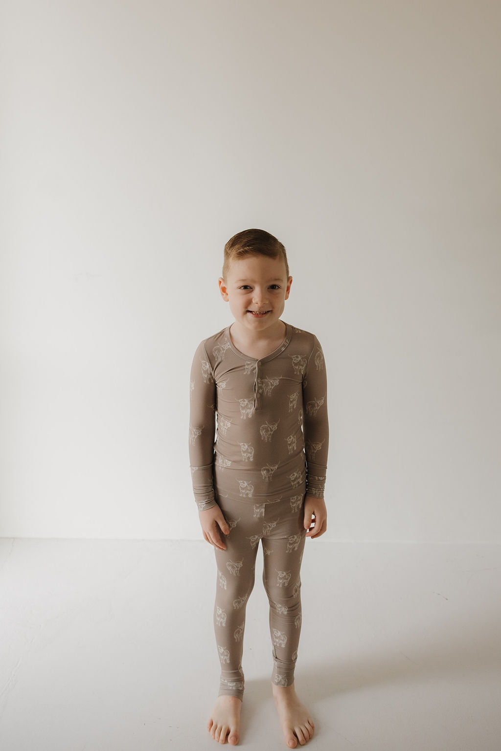 A young boy with short light brown hair stands barefoot on a plain white floor, smiling at the camera. He's wearing forever french baby's Bamboo Two Piece Pajamas | Highland with a white animal print pattern. The background is simple and white.