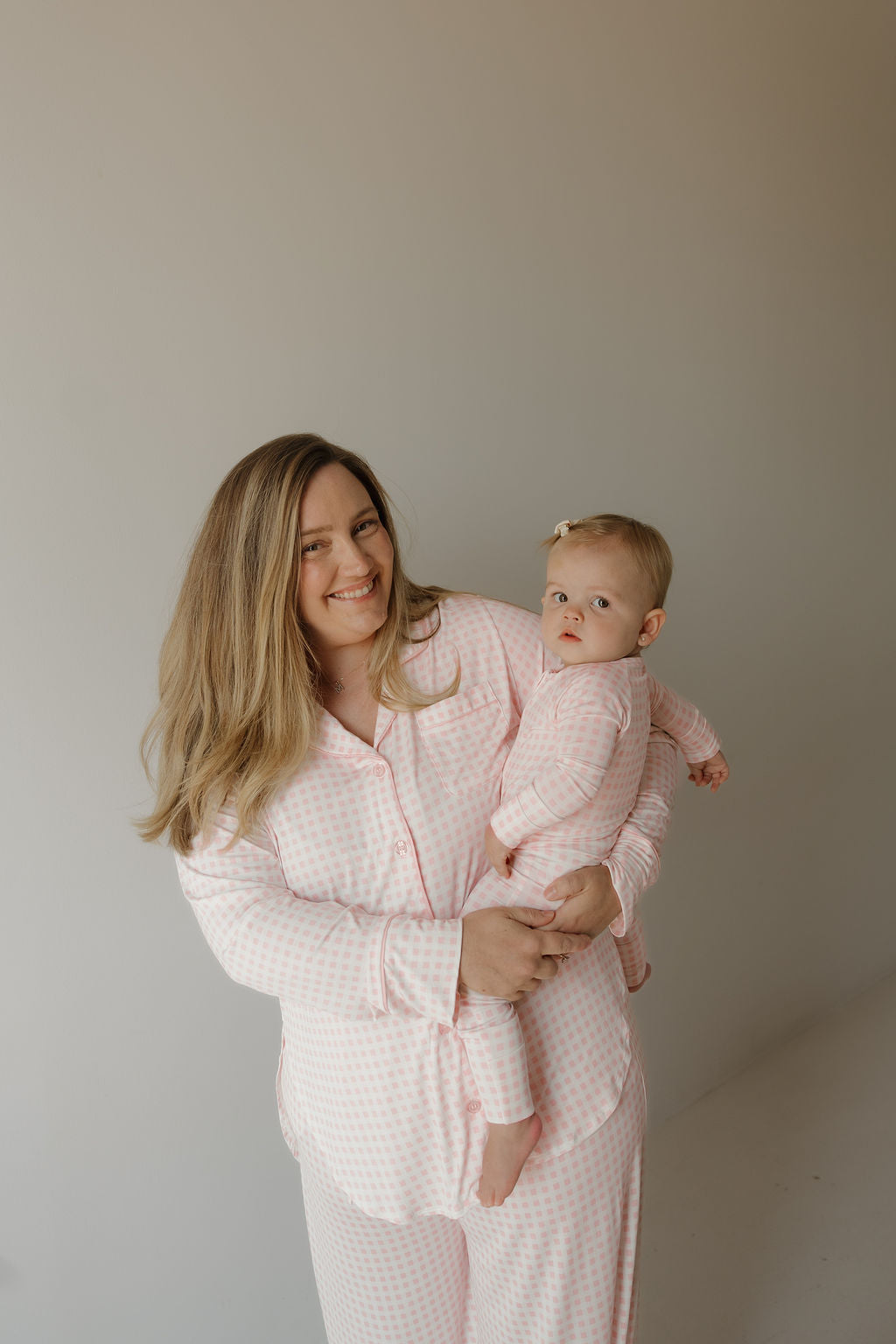 A woman and a baby, both dressed in matching forever french baby Giselle Women’s Bamboo Button Up Lounge Sets in pink and white checks, stand against a plain light-colored background. The woman is smiling as she holds the baby, who looks at the camera—highlighting the comfort and style of these hypo-allergenic loungewear sets designed for women and little ones alike.