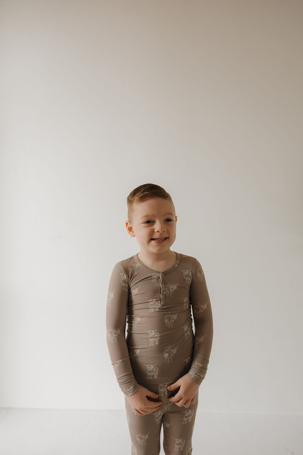 A smiling young boy with short light brown hair stands against a white wall, wearing forever french baby's Bamboo Two Piece Pajamas | Highland—a taupe, long-sleeve bamboo sleepwear set with a subtle animal print for cozy comfort.