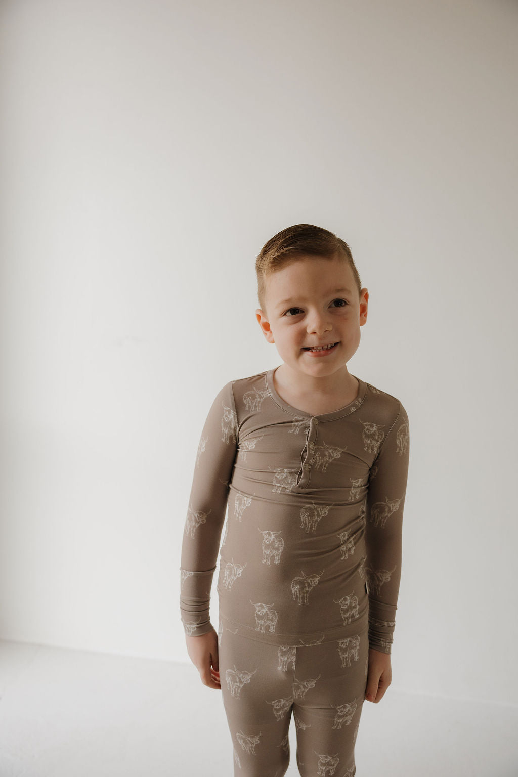 A young boy with short light brown hair smiles in forever french baby Bamboo Two Piece Pajamas | Highland, featuring a white animal print and hypoallergenic comfort, standing against a plain white background.