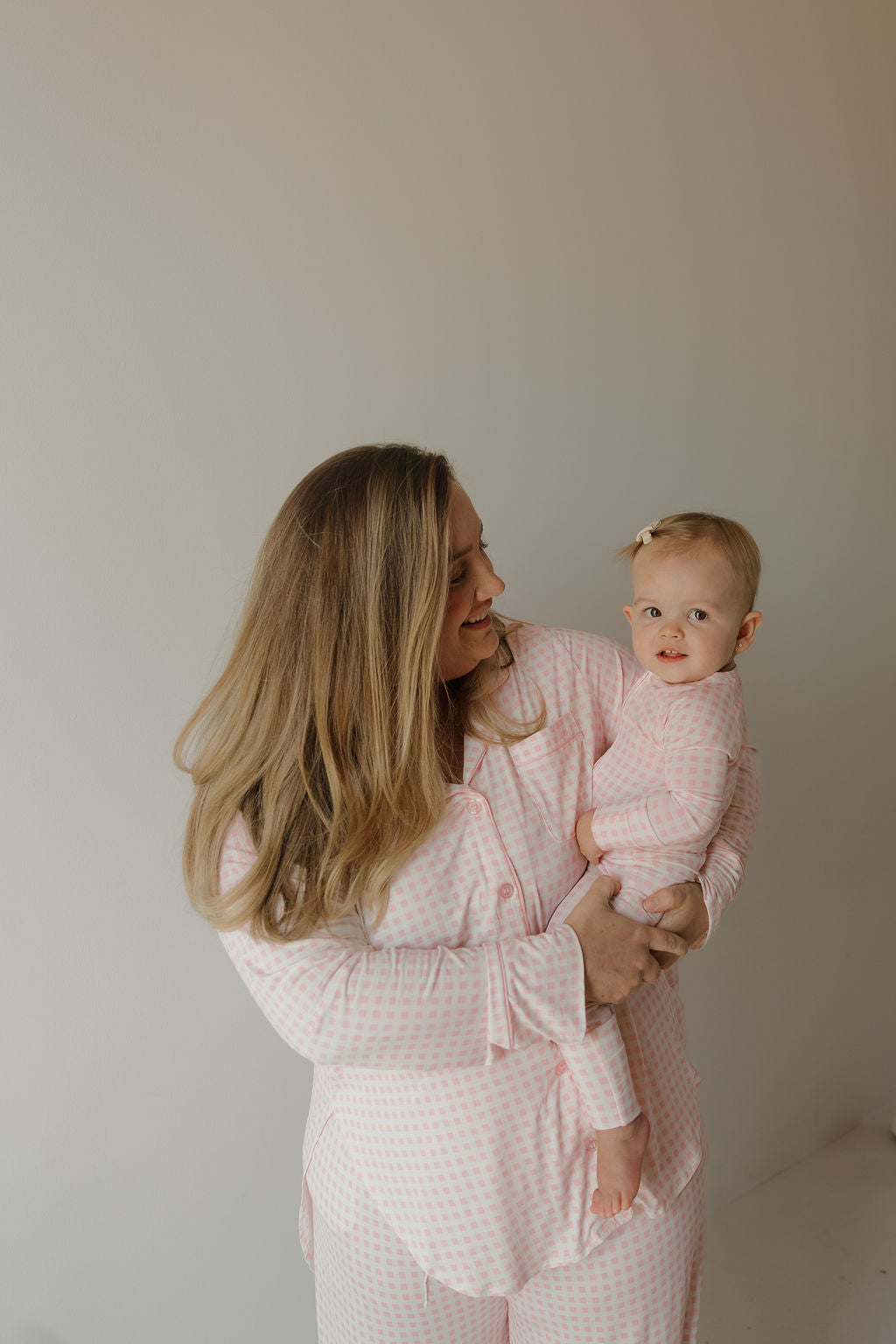 A woman with long blonde hair holds a baby, and both are wearing matching forever french baby Bamboo Zip Pajamas in the Giselle pink checkered pattern, standing against a plain light-colored wall.