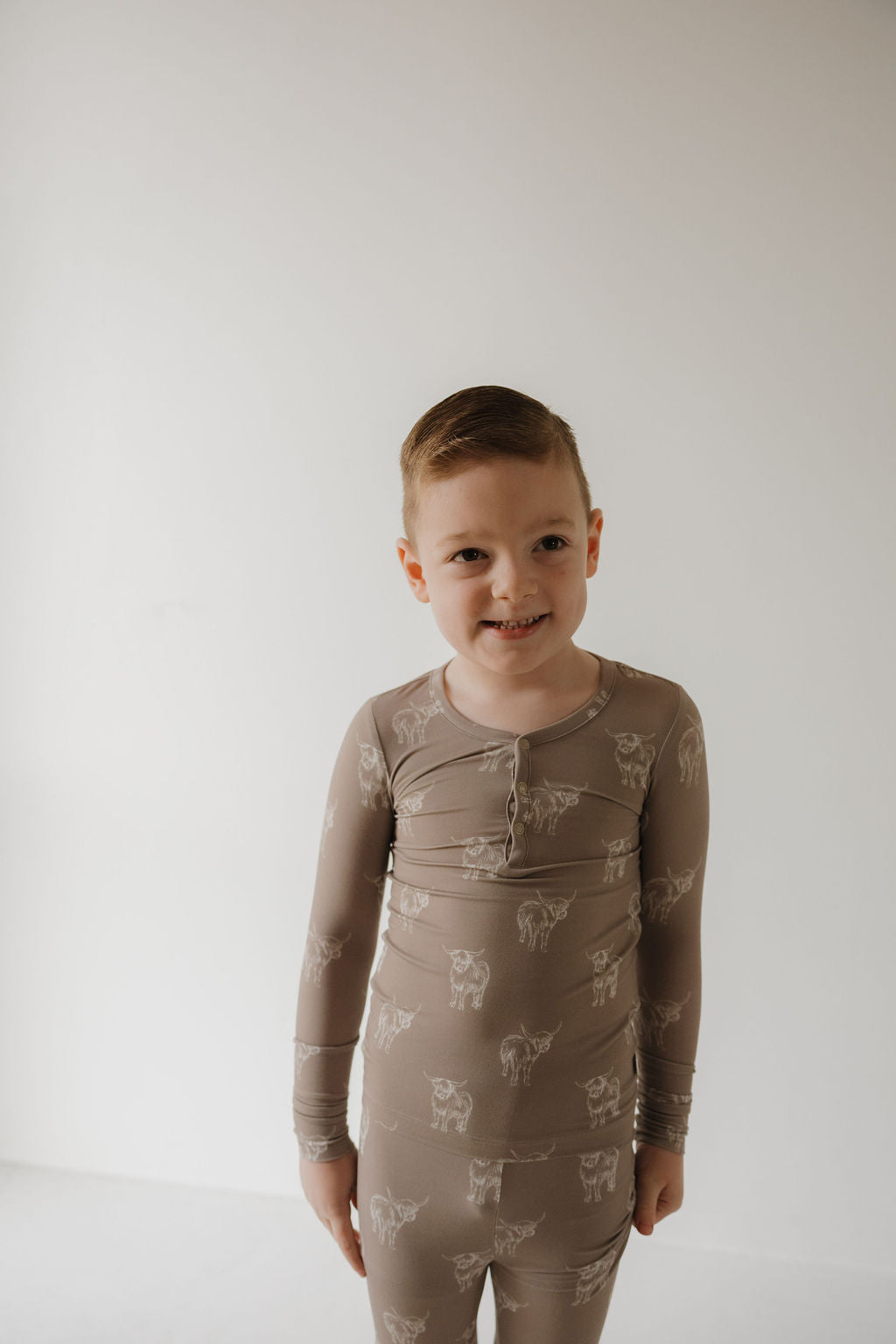 A young child with short brown hair smiles against a white background, wearing forever french baby’s Bamboo Two Piece Pajamas | Highland—beige, cow-printed, and hypoallergenic for cozy comfort.