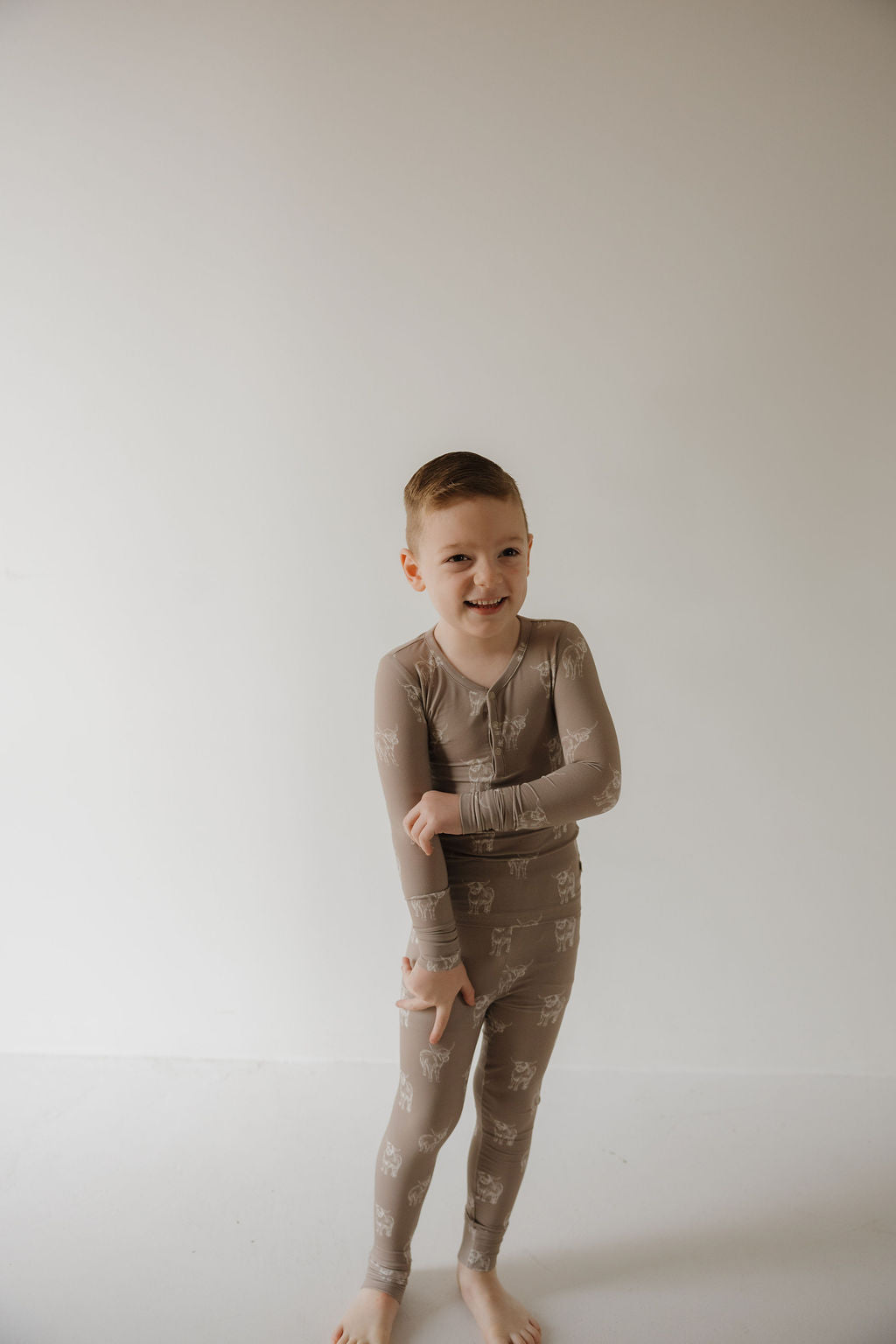 A young child stands barefoot against a white background, smiling and pointing down while wearing forever french baby’s Bamboo Two Piece Pajamas | Highland—cozy, hypoallergenic sleepwear ideal for sensitive skin.