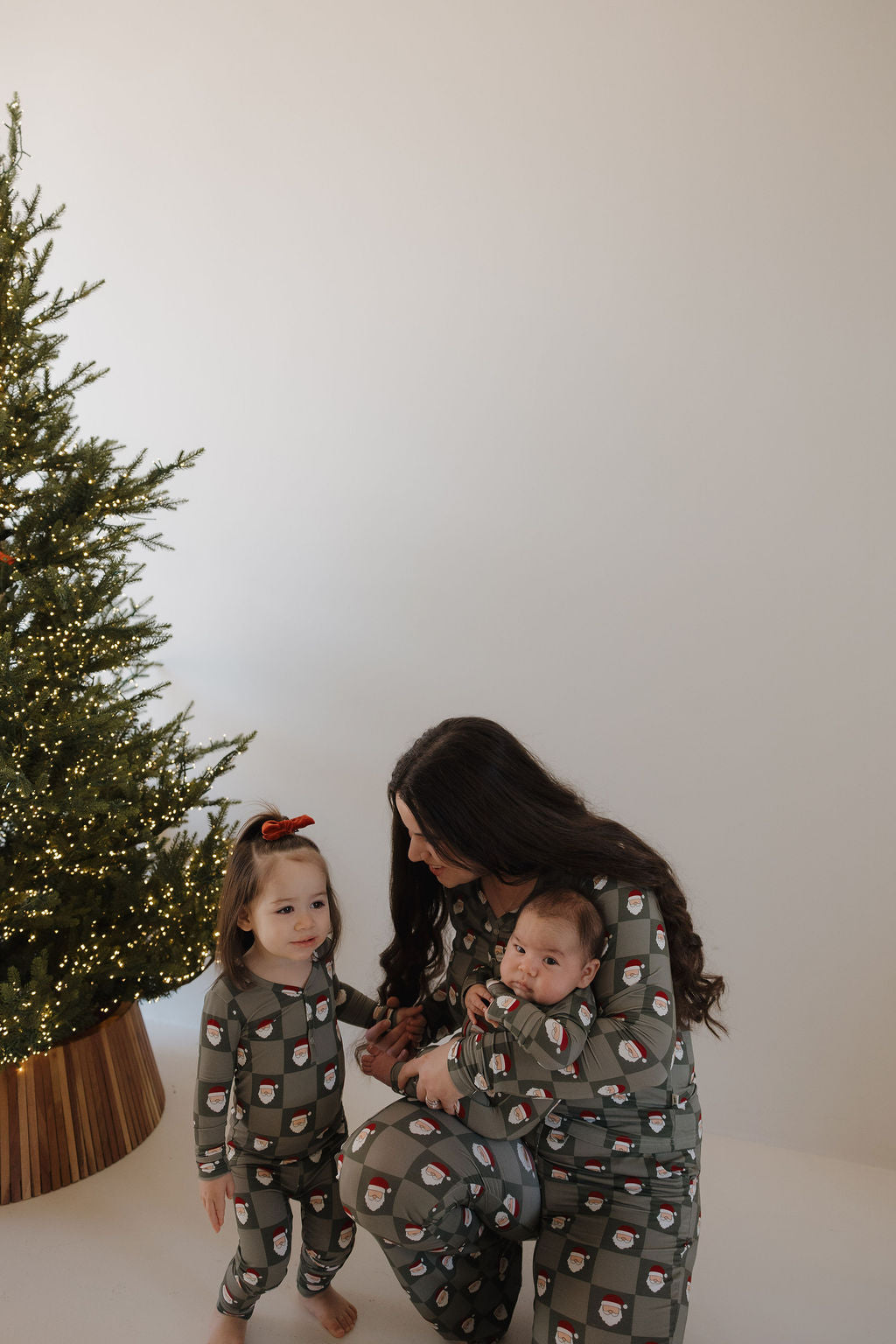 A woman and two children in forever french baby Bamboo Zip Pajamas | Santa Checkerboard sit by a lit Christmas tree. The woman holds an infant, both kids wear matching pajamas, the older one has a red bow, all on a white background.
