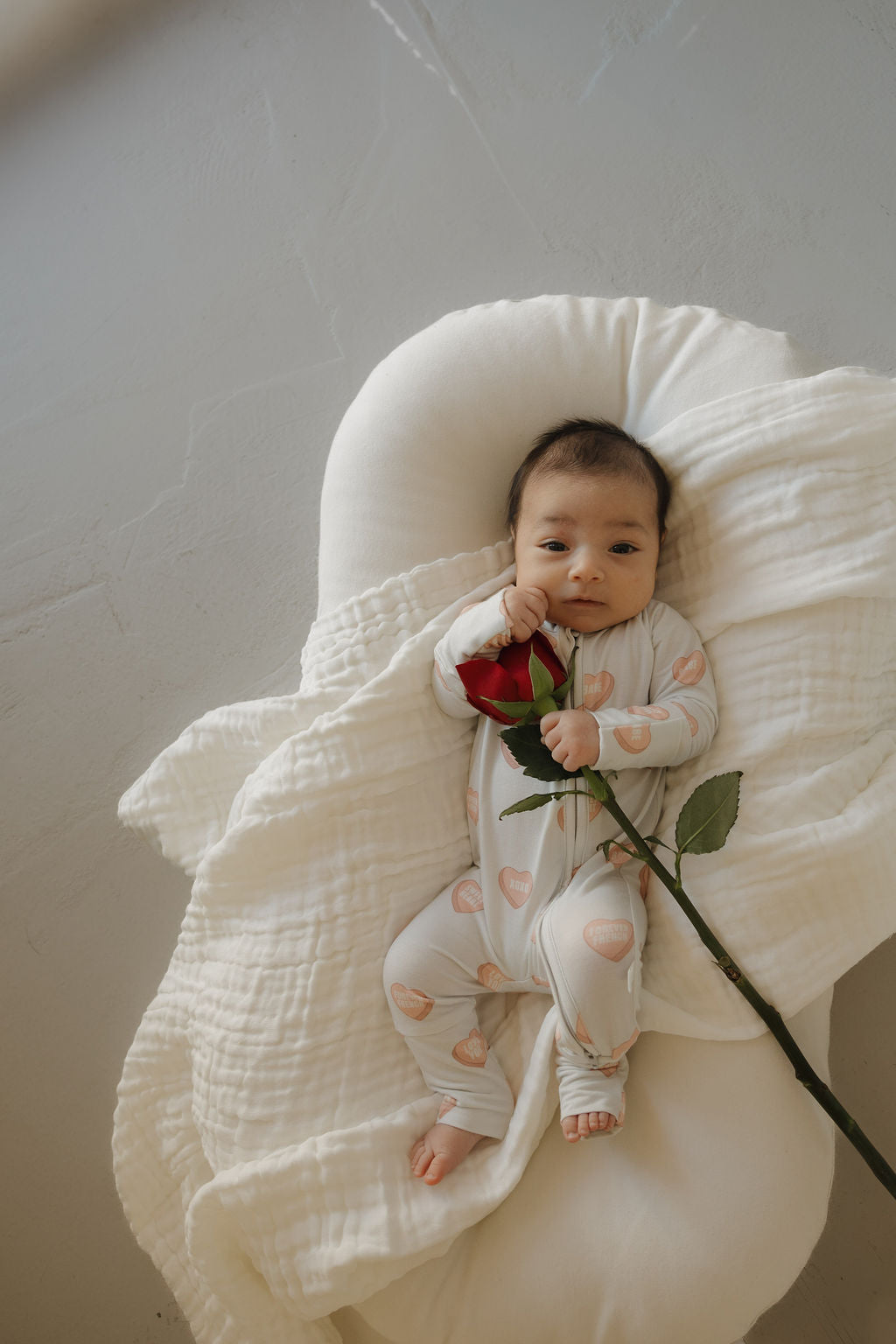 A baby in forever french baby's Bamboo Zip Pajamas | Love Notes - Grey with heart patterns lies on a white blanket, gazing upward while holding a red rose.