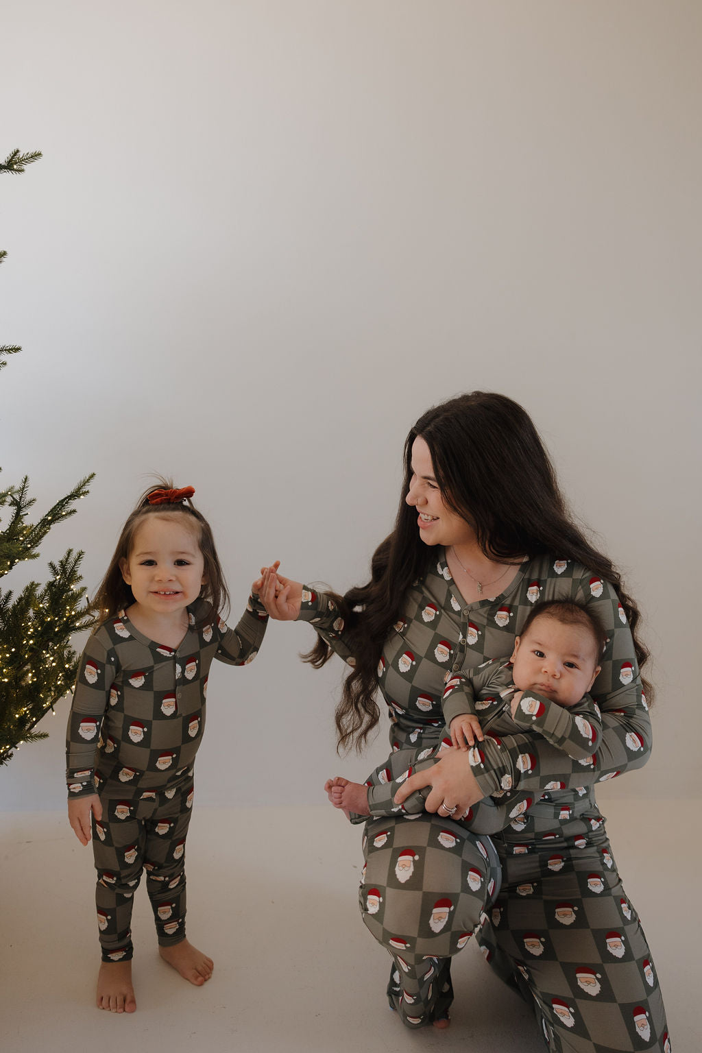 A woman and two children wear forever french baby Bamboo Zip Pajamas | Santa Checkerboard in green. The baby’s cozy, hypoallergenic pajamas match the little girl’s. A decorated Christmas tree is visible on the left.