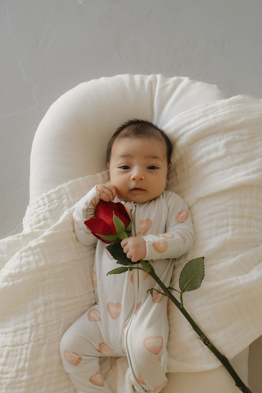 A baby in forever french baby Bamboo Zip Pajamas | Love Notes - Grey with heart patterns lies on a white blanket, holding a red rose and looking up at the camera.