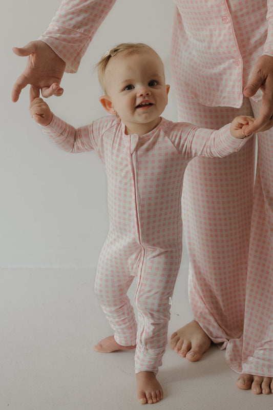 A smiling baby dressed in forever french baby’s Bamboo Zip Pajamas | Giselle—featuring a pink and white checkered bamboo fabric—stands barefoot on a light floor, holding an adult’s hands for support; both wear matching, breathable sleepwear.