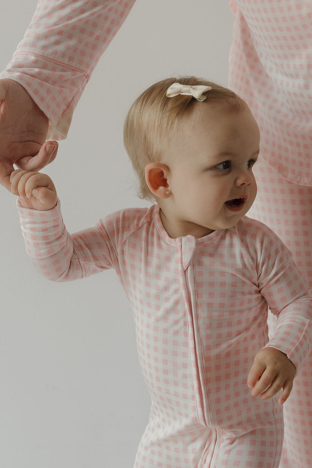 A baby wearing the Bamboo Zip Pajamas | Giselle by forever french baby—crafted from hypoallergenic bamboo fabric in a pink and white checkered pattern—holds an adult’s hand. The baby has a small bow in her hair and is looking to the side, smiling.