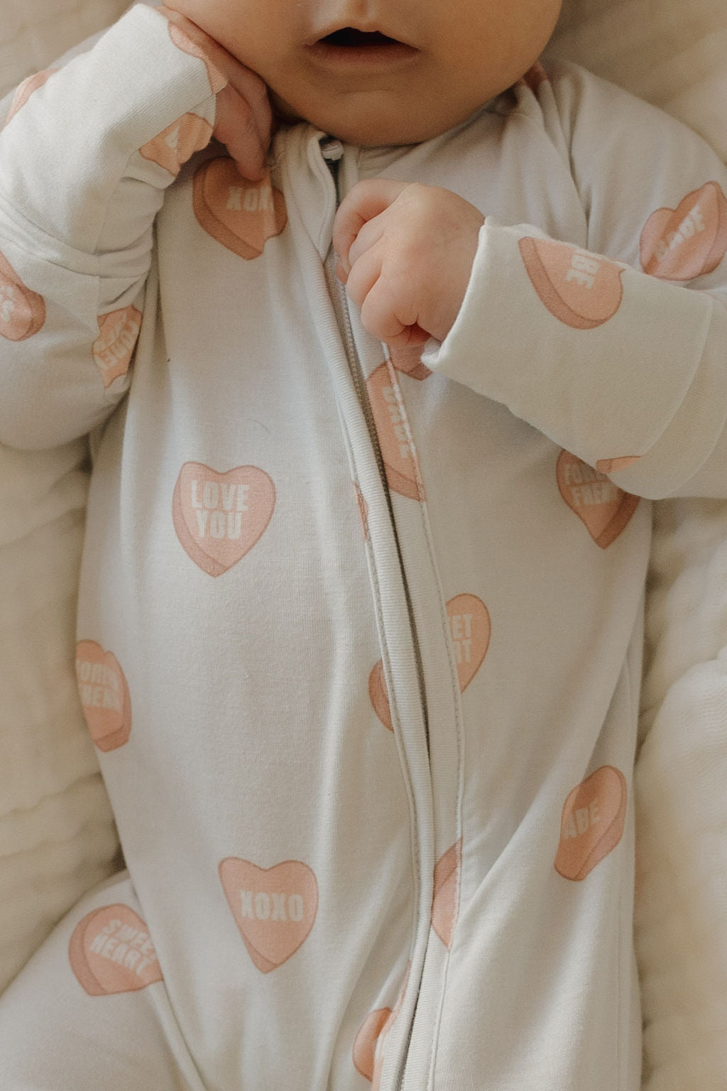 A baby in forever french baby's Bamboo Zip Pajamas | Love Notes - Grey, featuring heart-shaped candies with "LOVE YOU," "XOXO," and "CUTIE" prints. The baby’s face is partly visible, one hand near their mouth.