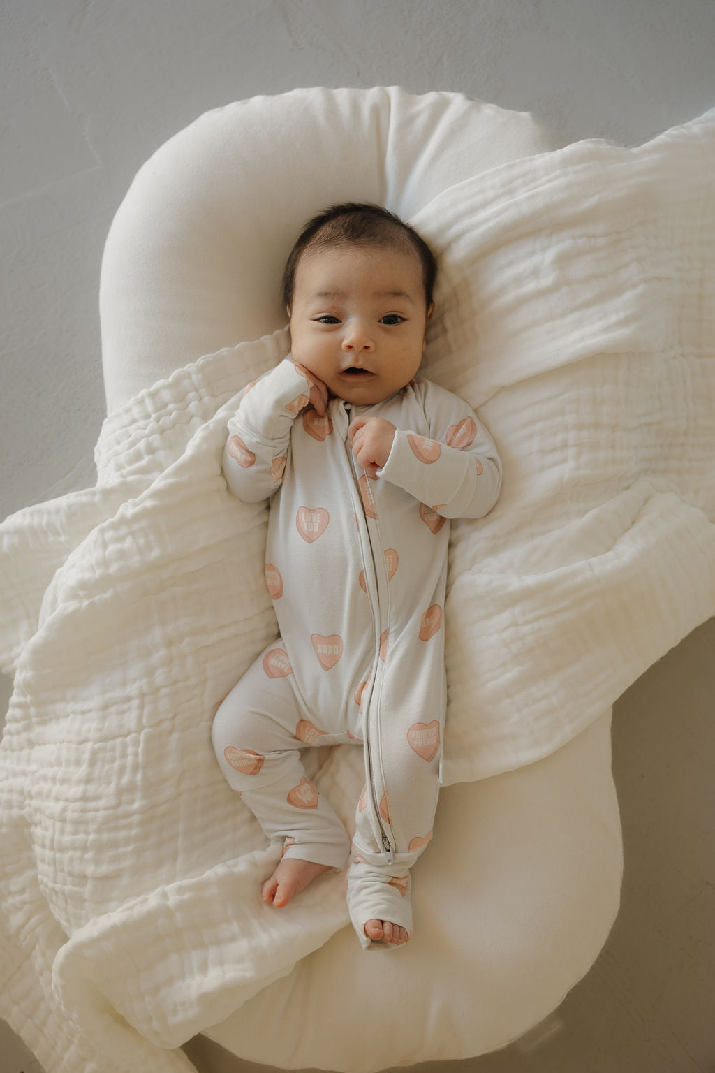 A baby lies on a soft white surface, wrapped in a blanket and wearing forever french baby's Bamboo Zip Pajamas | Love Notes - Grey, a breathable onesie. The baby looks up with one hand near their face.