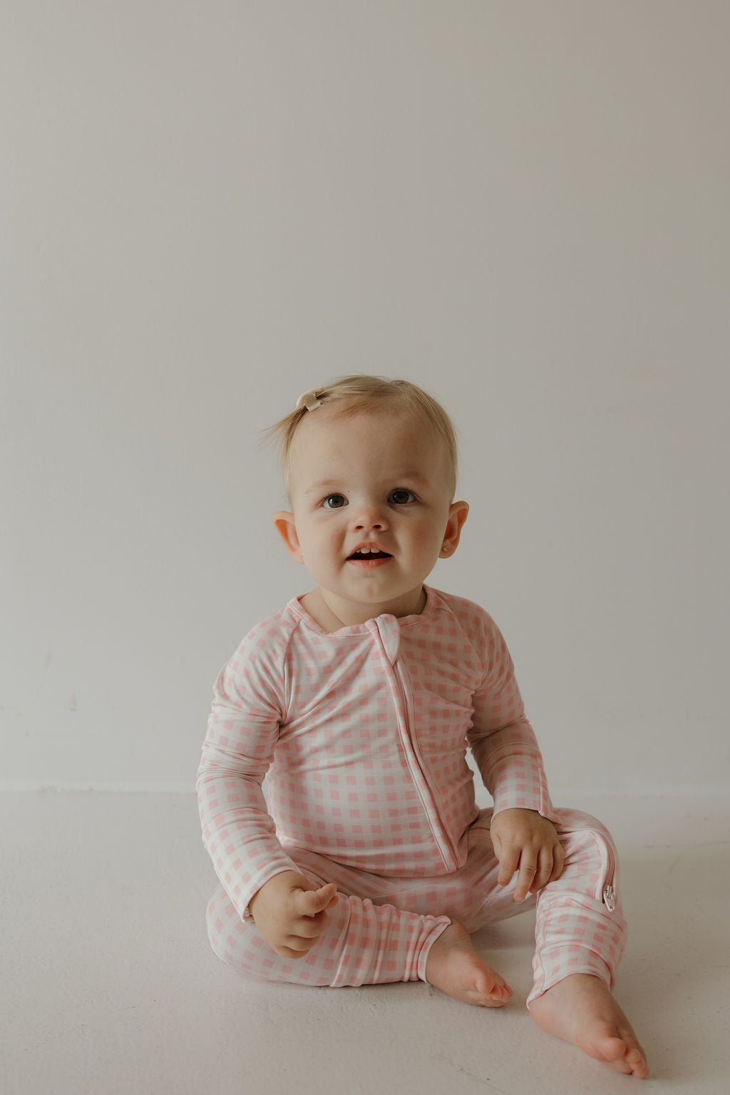 A light-haired baby with a hair clip sits on the floor against a white background, smiling gently and looking slightly up while wearing the forever french baby Bamboo Zip Pajamas | Giselle—a pink and white checkered, hypoallergenic outfit.