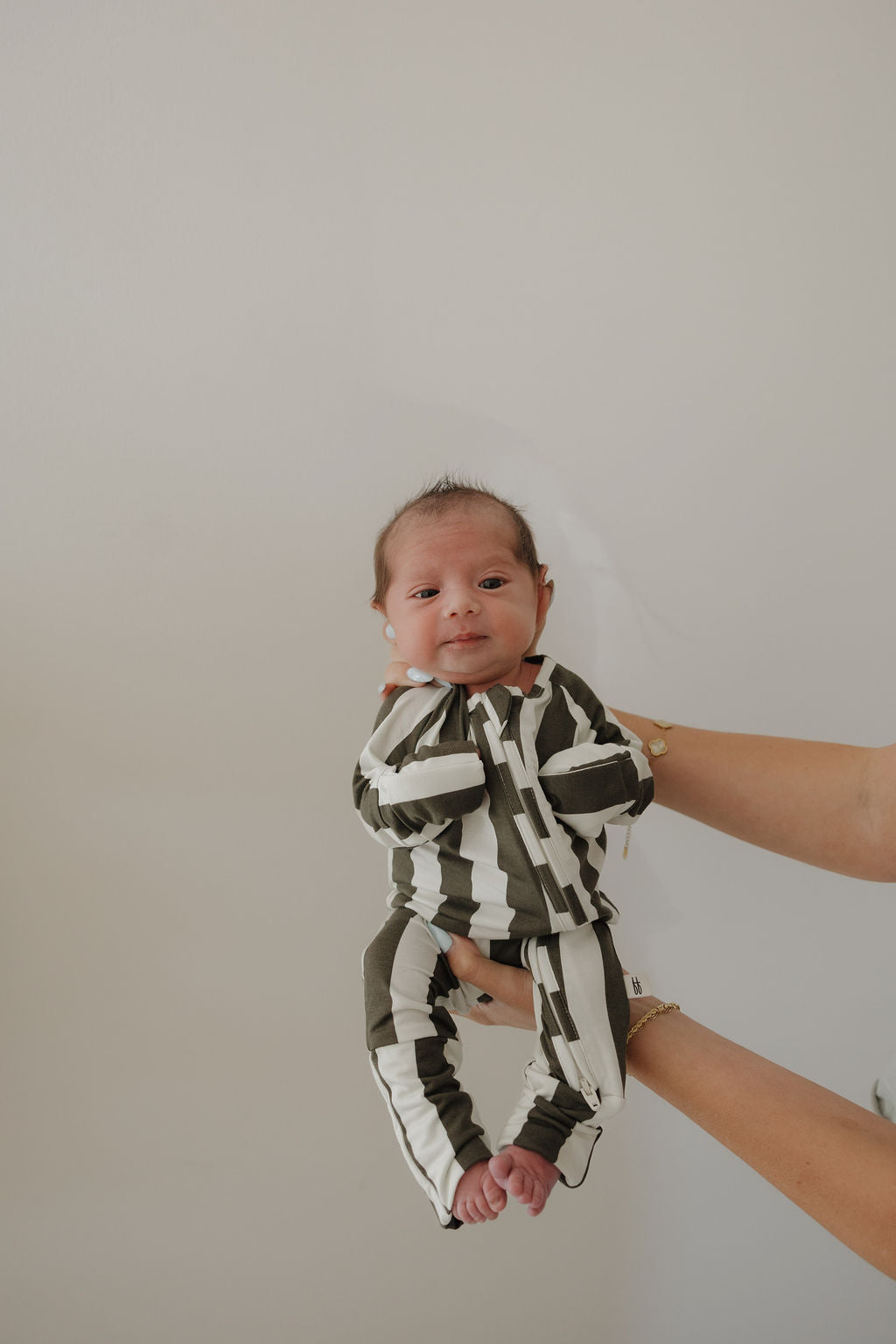 A baby in forever french baby Bamboo Zip Pajamas | Fireside Stripe is held up by adult hands against a plain background, looking calmly at the camera in these breathable green and white striped pajamas.