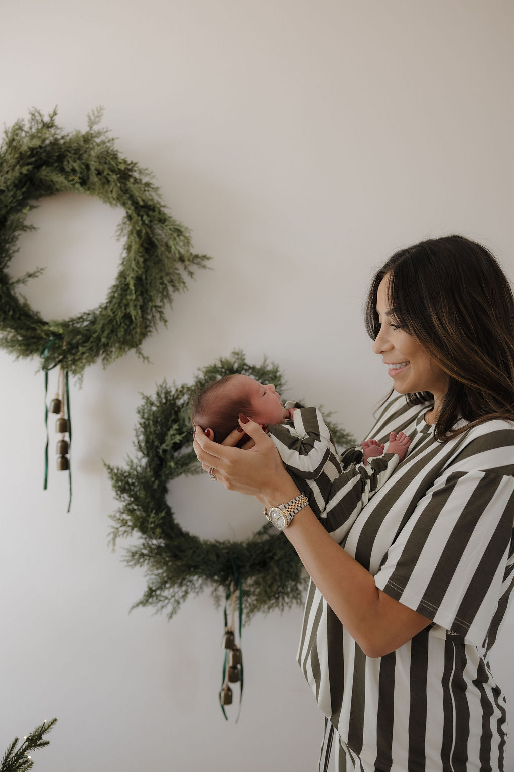 A woman wears forever french baby Bamboo Zip Pajamas in Fireside Stripe, holding her smiling newborn in matching sleepwear. Behind them, two green wreaths with bells and ribbon hang on a white wall.