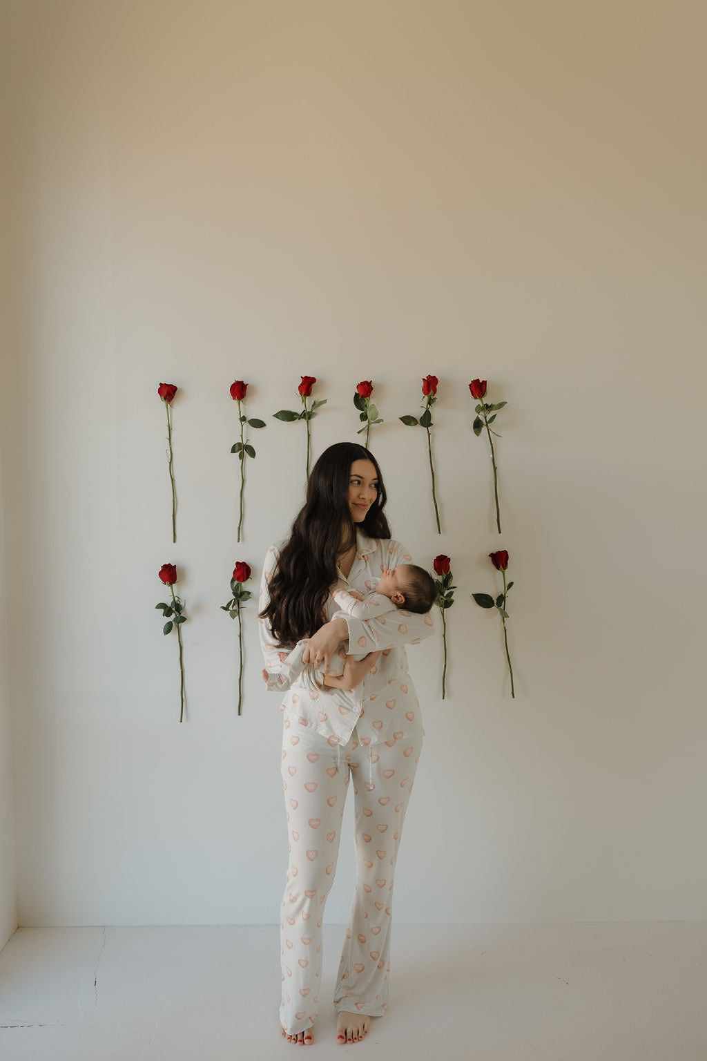 A woman in the forever french baby Women's Bamboo Button Up Lounge Set | Love Notes - Grey holds a baby, both barefoot by a white wall with nine red roses—enjoy everyday comfort and style in hypoallergenic bamboo clothing.