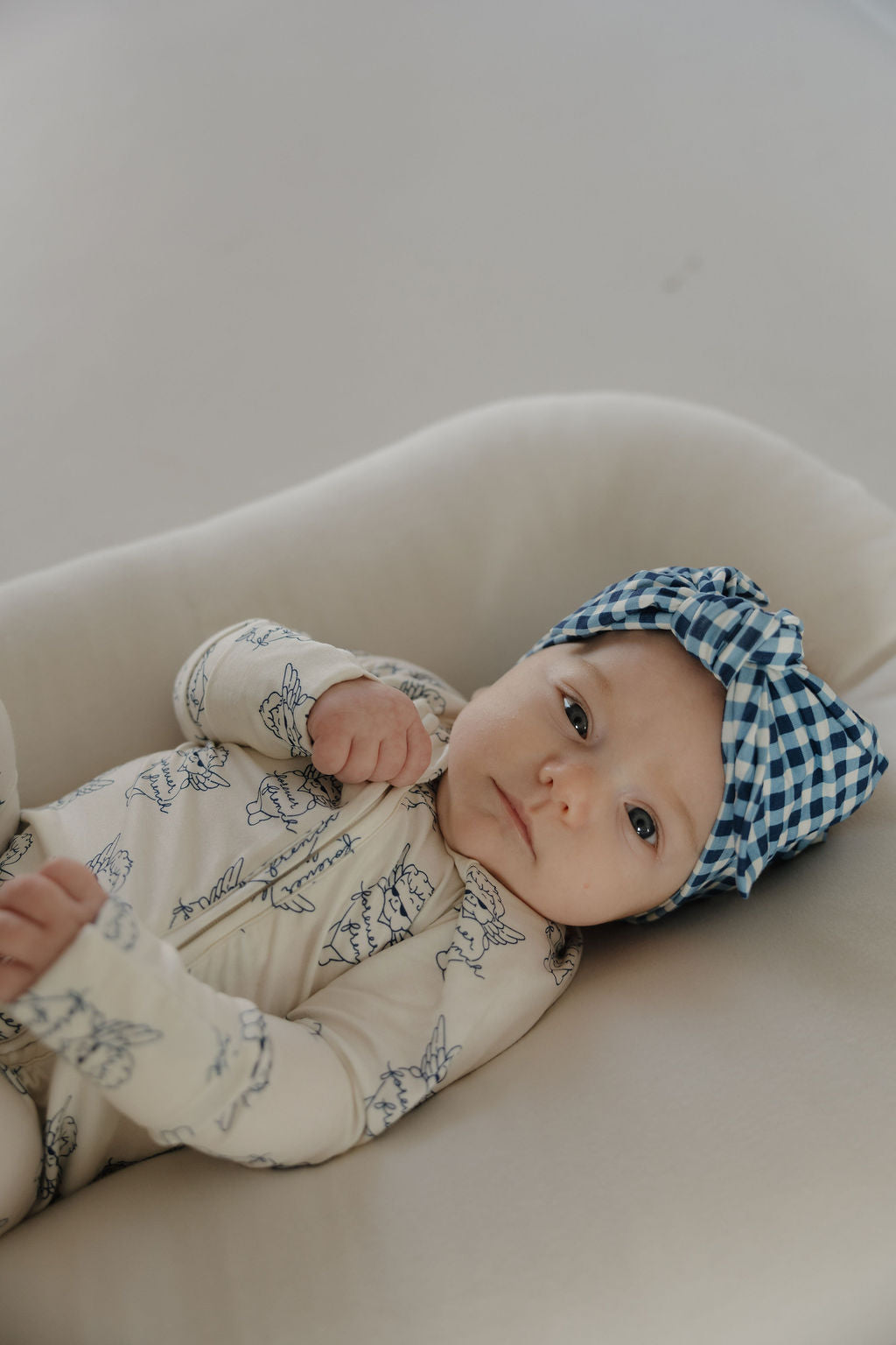 A baby wearing the forever french baby Bamboo Head Wrap in Blue Gingham lies on a soft, light-colored cushion and looks up at the camera.