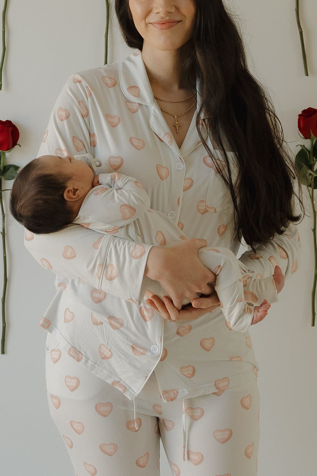 A woman in forever french baby Bamboo Zip Pajamas | Love Notes - Grey holds a sleeping baby in matching hypo-allergenic pajamas. Both stand by a white wall decorated with two red roses. The woman, with long brown hair, smiles gently.